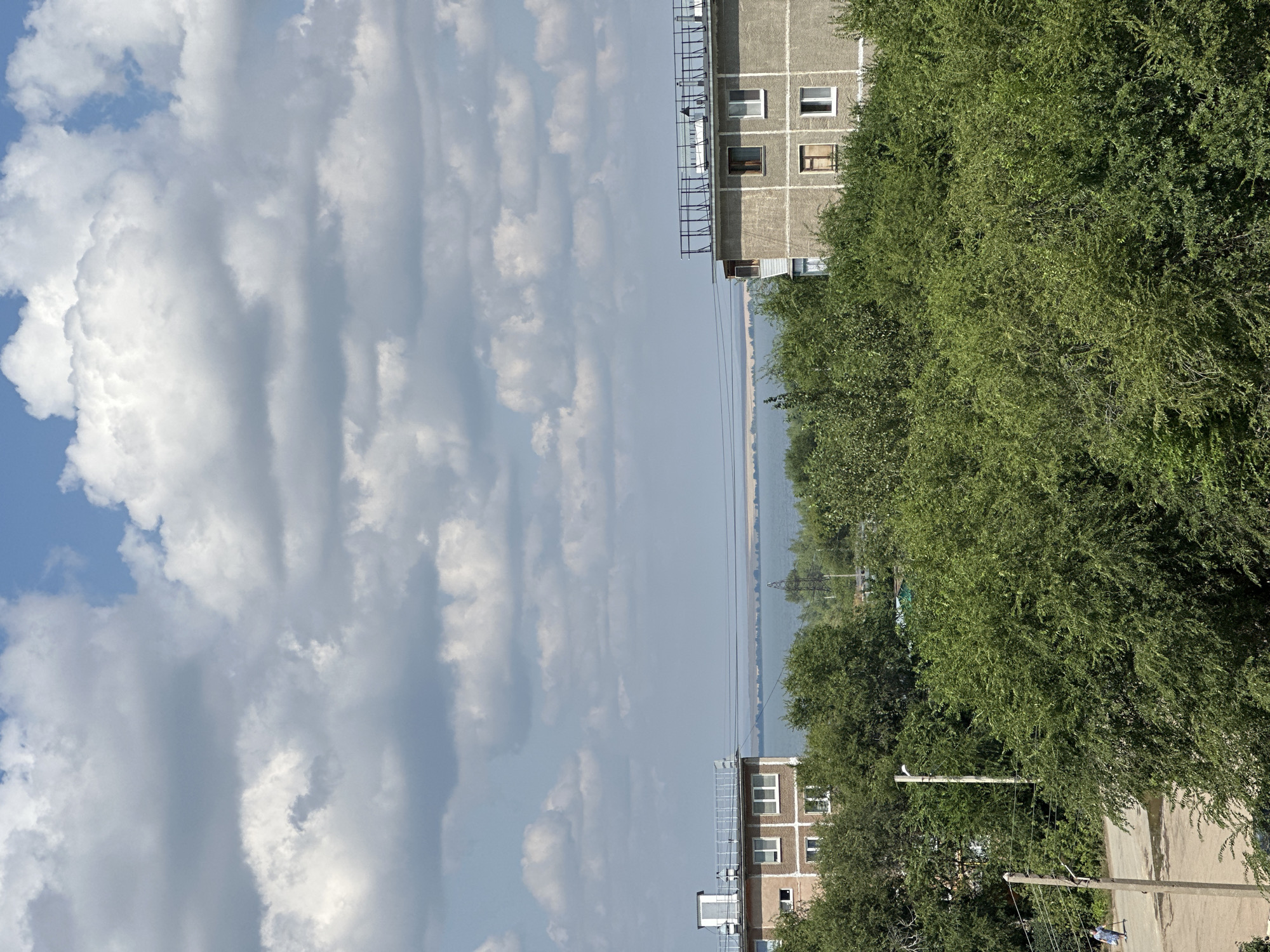 The image captures a view of the city of Energetik, Russia, on a cloudy day. The foreground is dominated by lush, green trees, obscuring much of the lower half of the scene. Behind the trees, two apartment buildings are visible. The building on the left is lighter in color and has a more traditional facade with visible windows and a rooftop structure. The building on the right is gray and appears to be a more utilitarian structure with balconies and evenly spaced windows. In the background, a wide body of water, likely the pond of the Nikolaevskaya thermal power plant, stretches across the horizon, bordered by a distant treeline and fields. Power lines are visible crisscrossing the landscape between the buildings and the water. The sky is filled with a mix of white and gray clouds, with patches of blue sky peeking through. A single person, a woman wearing a blue dress, is walking on a paved area on the left side of the image, near a utility pole.