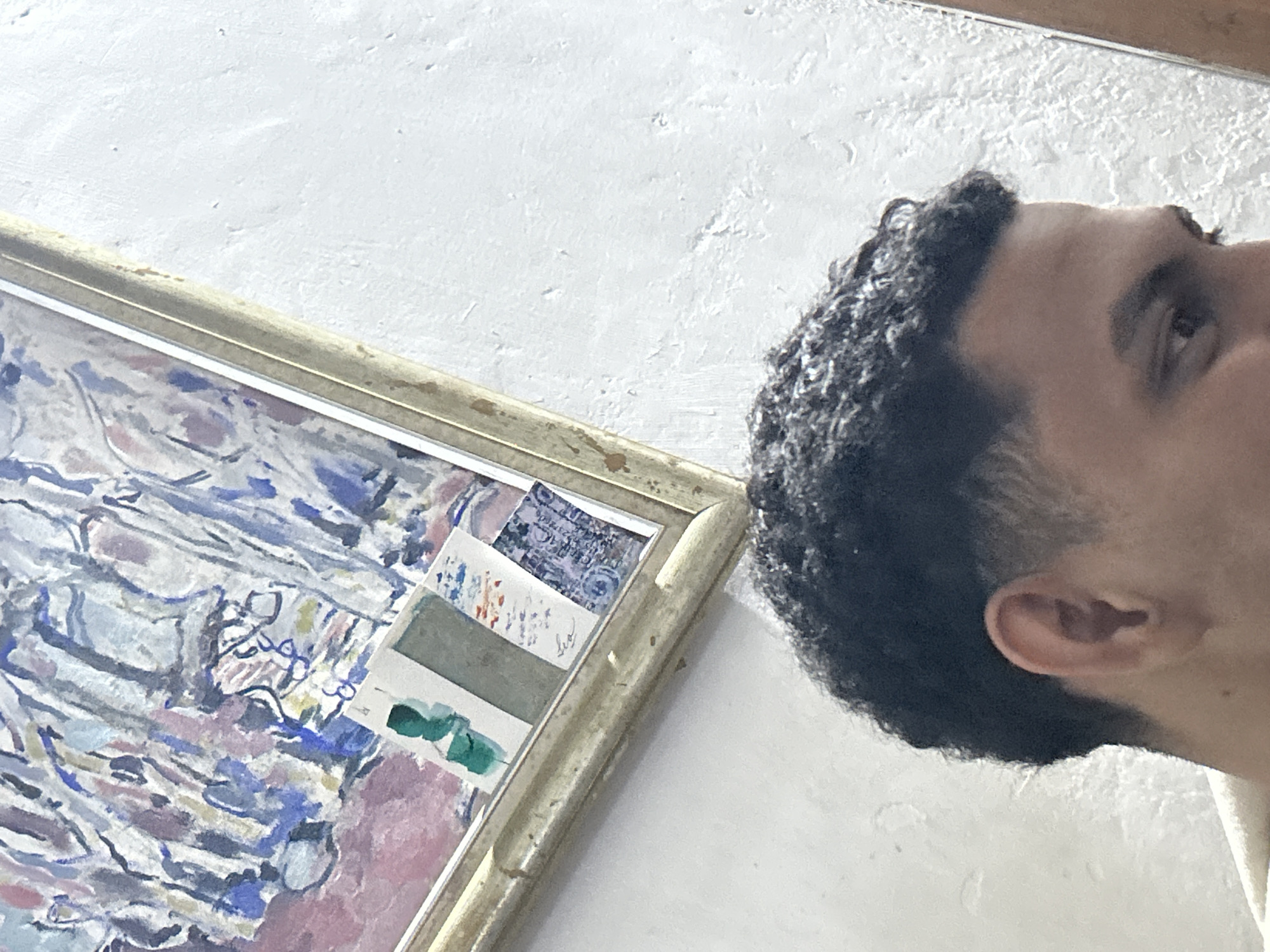 The image, captured in Santa Ana, Venezuela, shows a close-up, angled view of the right side of a person's head, from just above the ear to the eyebrow. The individual has dark, curly hair with a faded haircut. In the background, a framed abstract painting hangs on a textured white wall. The painting features a vibrant palette of blues, pinks, and purples with visible brushstrokes. The frame of the painting appears to be made of a light-colored, possibly gilded material. A few small, rectangular cards or prints are adhered to the lower portion of the painting, some displaying colors like green and blue, and one showing intricate patterns. The overall lighting suggests an indoor setting.