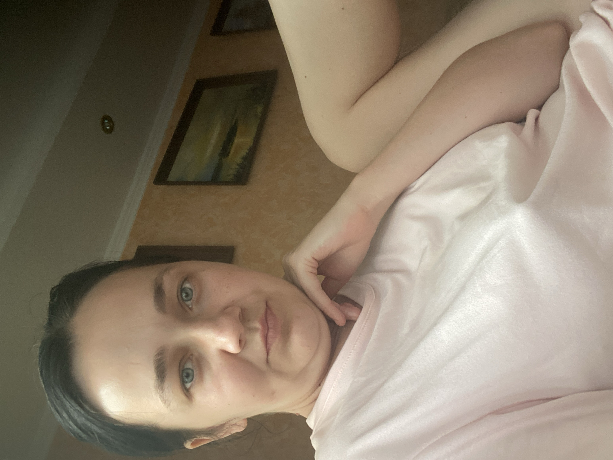 A medium shot taken from a slightly low angle shows a young woman with dark hair and light blue eyes looking directly at the camera. She is wearing a pale pink t-shirt and resting her chin on her right hand, which is positioned on her chest. Her left arm is bent and rests on her body, partially obscured by the t-shirt. Her skin is fair with a few freckles visible on her nose and cheeks. The background consists of a light-colored wall with two framed paintings. One painting, to the right, depicts a landscape with a body of water and a setting sun. The ceiling is visible at the top, with a recessed light fixture. The overall lighting suggests an indoor environment.