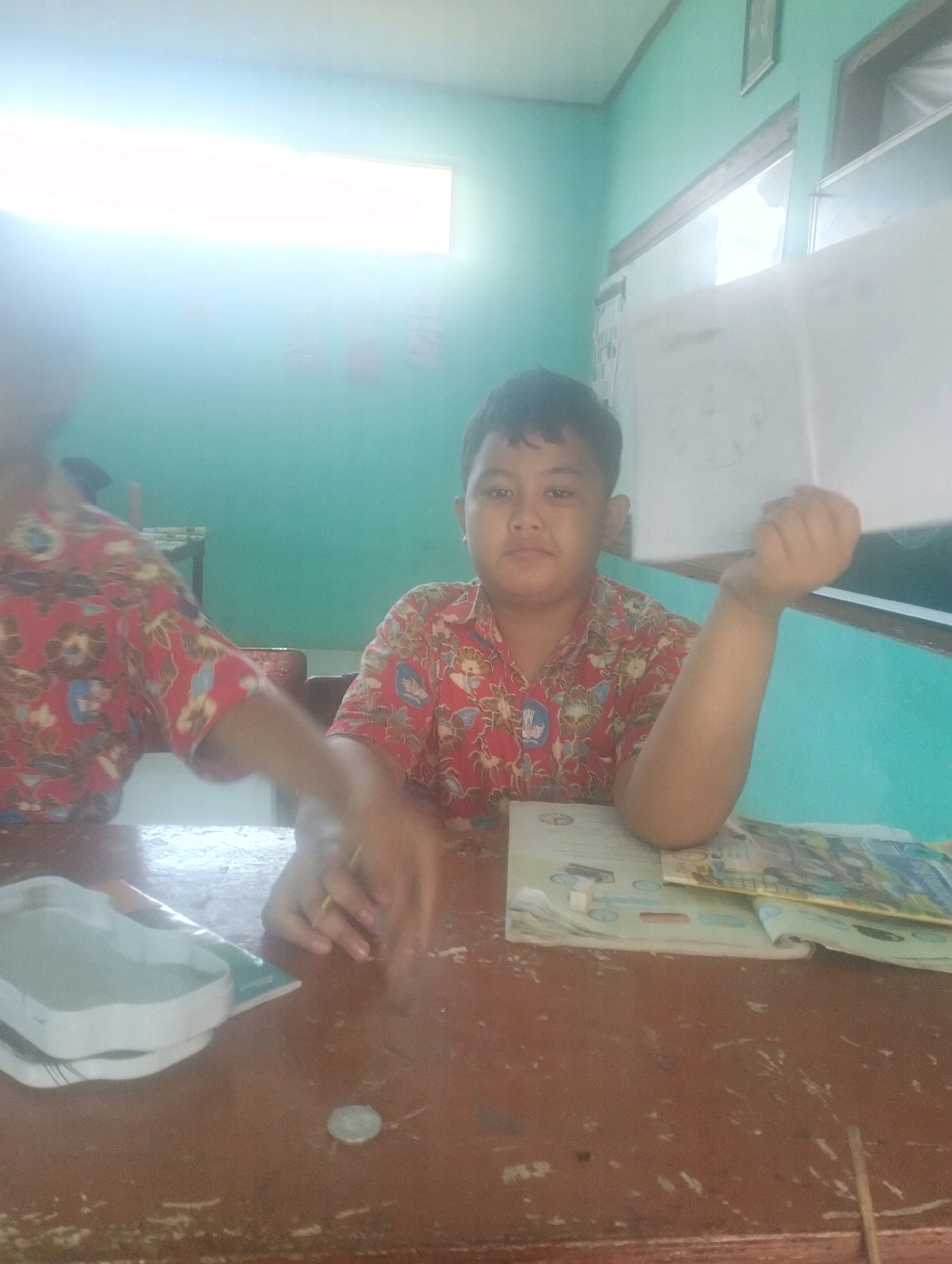 In a classroom in Blabakan, Indonesia, two young boys sit at a wooden desk. The boy in the foreground, on the right, wears a red shirt with a white floral pattern and is looking at the camera. His right arm is raised, with his hand clenched into a fist. On the desk in front of him are several books. The boy on the left, partially obscured, also wears a similar red floral shirt and appears to be interacting with something on the desk. The room has teal-colored walls and a large window with a bright light source, possibly fluorescent tubes, above it.