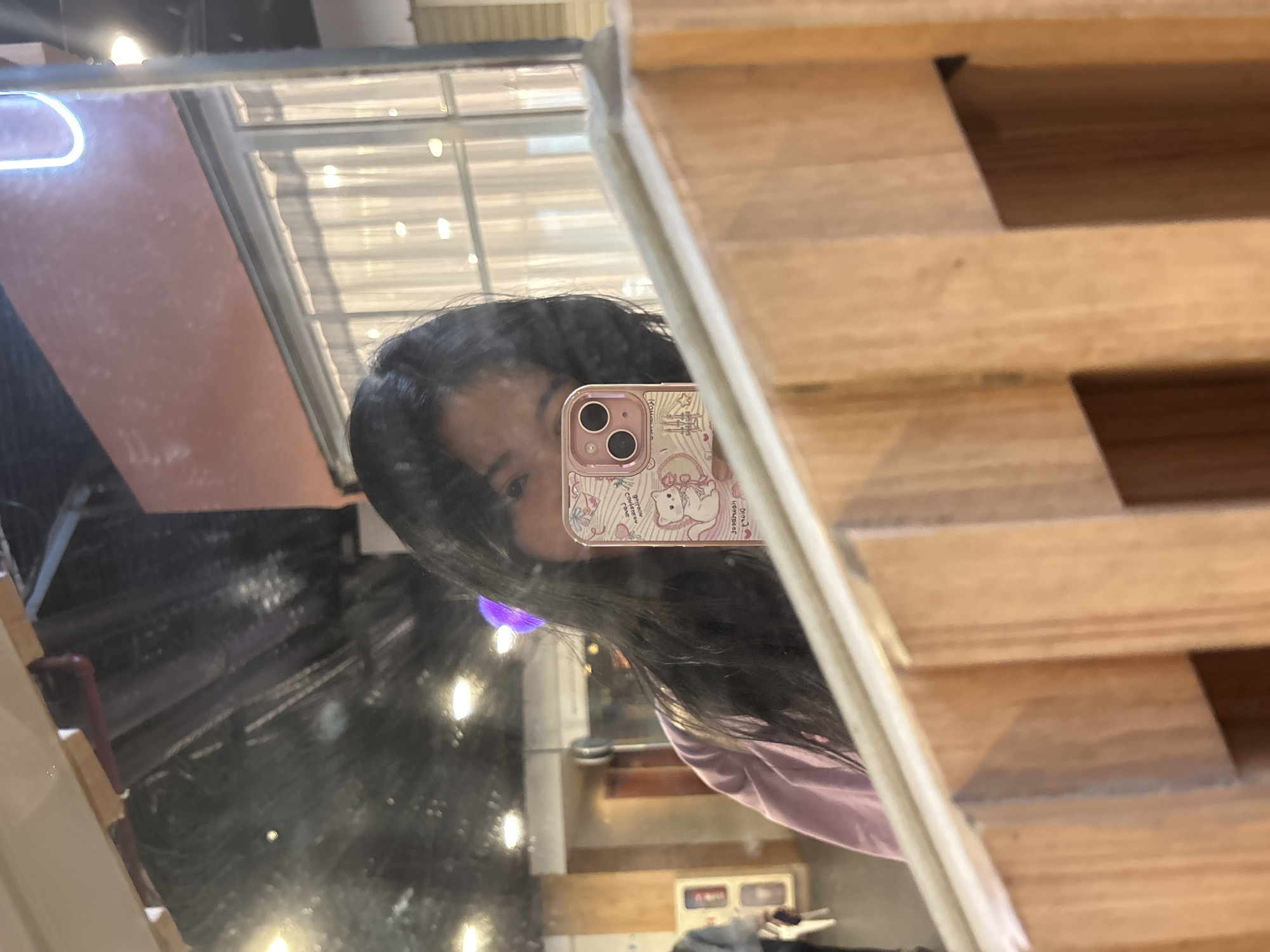 A person is reflected in a mirror, holding up a phone with a patterned case in front of their face. The mirror reveals a partial view of an indoor space with a glimpse of what appears to be commercial or industrial equipment. Wooden structures partially obstruct the reflection. The image's location is not discernible from the visual information provided.