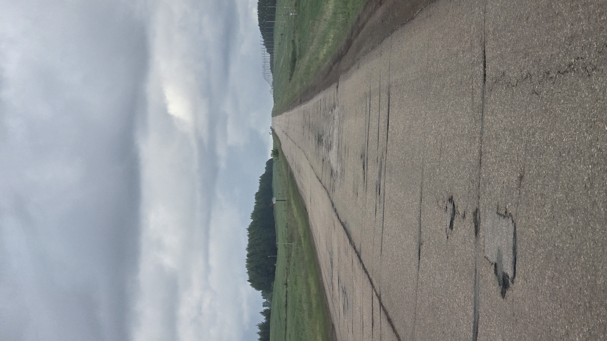 The image, captured near Солояз, Russia, depicts a long, straight, two-lane asphalt road extending into the distance under a heavily overcast sky. The sky is dominated by layered, grey clouds, indicating diffuse, flat lighting with no direct sunlight.
The asphalt road surface is significantly deteriorated, characterized by extensive longitudinal and transverse cracks, alligator cracking, and numerous distinct potholes varying in size and depth, particularly concentrated in the center of the lanes. Some dark patches on the road surface suggest moisture or recent repairs.
Green fields flank both sides of the road, ascending gently toward the horizon. To the left, a dense line of dark green coniferous trees forms a natural boundary against the sky. A small white sign with faded red text, possibly indicating a distance marker like "17KM," is visible on the left shoulder approximately midway down the visible road.
On the right side, a series of slender utility poles with horizontal cross-arms extends linearly into the distance, paralleling the road. The road itself recedes towards a crest in the far distance, obscuring anything beyond.
No individuals, vehicles, or animals are present in the scene, rendering it desolate. The overall color palette is muted, consisting primarily of various shades of grey from the sky and road, and dark greens from the vegetation. The perspective is low, looking directly down the road toward the distant horizon.