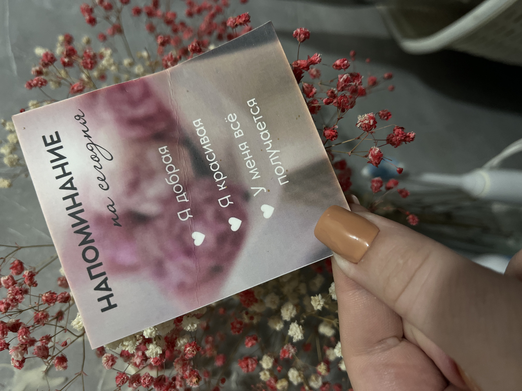 A close-up shot shows a person holding a small card with Russian text. The card's background features a soft pink hue, likely from a floral image. The text on the card reads: "НАПОМИНАНИЕ на сегодня. Я добрая. Я красивая. У меня всё получается," interspersed with small heart icons. A person is holding the card with their right hand, with a single finger visible with a tan-colored nail polish. The card is surrounded by small, dried red and white flowers. The environment is blurred but appears to be indoors. There is no indication of the city of Chaikovsky, Russia.