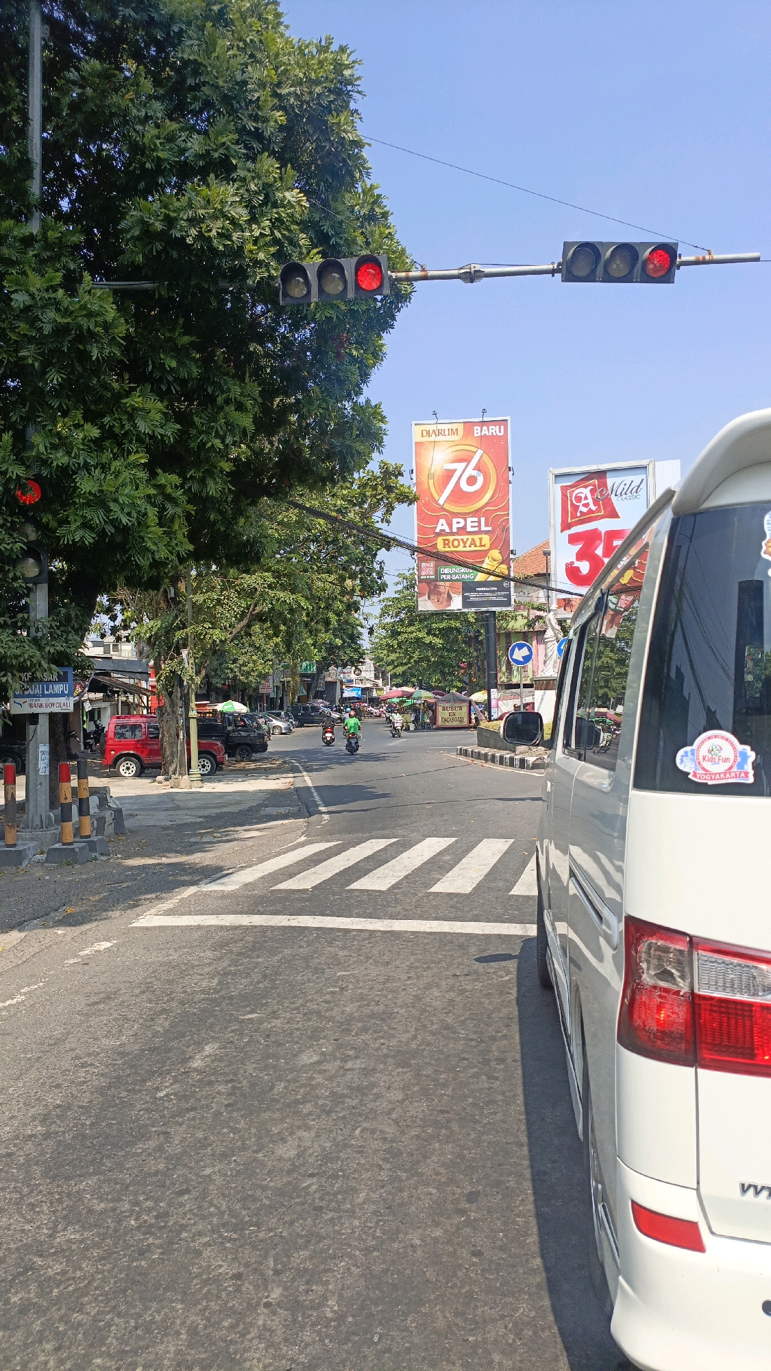 The image shows a street scene in Sidoharjo, Indonesia, on a sunny day. A white van is parked on the right side of the road, facing away from the viewer. In the foreground, a crosswalk is visible, with white stripes on the asphalt. To the left of the road, a large green tree with dense foliage is present, partially obscuring a traffic light. The traffic light is red. Further down the street, there are several shops and stalls, with people and vehicles, including motorcycles and cars. Two large billboards are visible in the background, one advertising "Djarum Baru 76 Apel Royal" and another for "A Mild Classic" with the number "35". The sky is clear and blue.