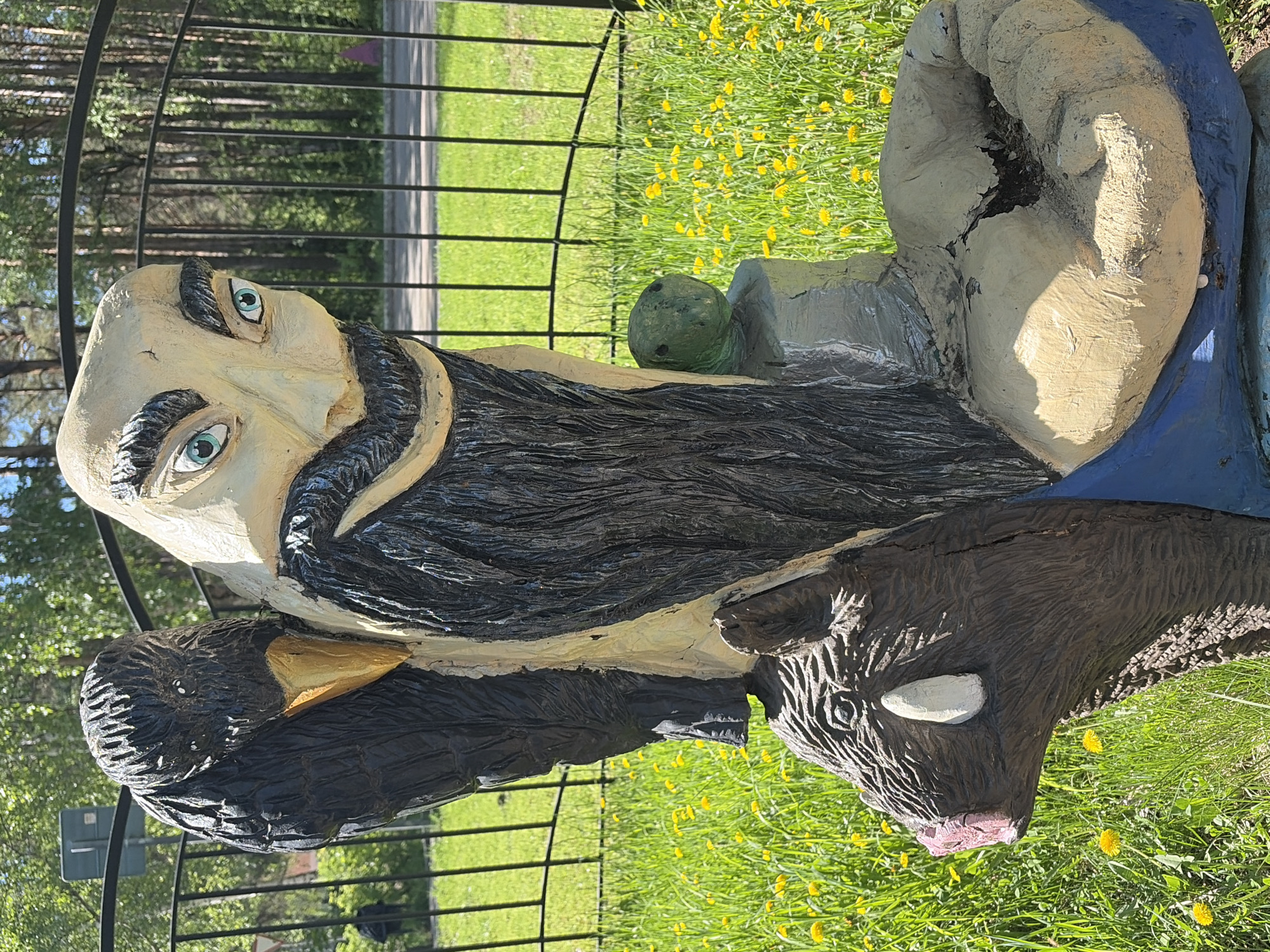 The image shows a painted, wooden sculpture of a man with a long, black beard. His eyes are painted blue. The sculpture also includes a black bird with a yellow beak, a brown bear, and a green turtle. The sculpture is situated on a grassy area covered in yellow dandelions, near a black metal fence. The background includes trees and a paved road. This statue is located in the city of Chaikovsky, Russia.