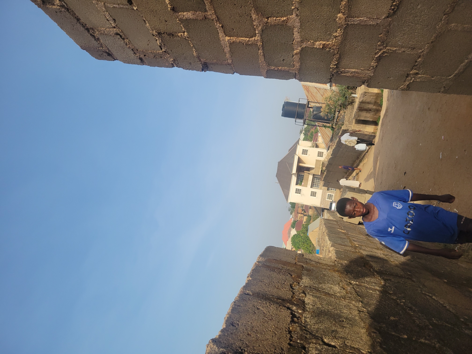 The media file is an image, rotated 90 degrees clockwise, depicting an outdoor scene in what appears to be a residential or semi-urban area in Jos, Nigeria.
In the corrected orientation, the image captures a view between two rough-hewn concrete block walls. On the right, a young boy stands on an unpaved ground surface, looking towards the camera. He wears a bright blue short-sleeved t-shirt with "HummEL" visible on the sleeve and "CANCUN" prominently displayed in white capital letters on the chest, along with other smaller, less legible text below it. He has dark hair and a neutral expression.
Beyond the boy and the wall on the right, an open area or alleyway extends, leading to several buildings in the background. An adult man in a white t-shirt is visible further back, walking and carrying two large white containers or bags. Another smaller figure can be seen in the distance near the buildings.
The structures in the background include a cream-colored building with multiple grid-patterned windows and other buildings with reddish, corrugated iron roofs. A black water tank on a metal stand is visible above one of the roofs, suggesting common water storage methods in the region. The ground is unpaved, consisting of dirt or gravel.
The sky is a clear, bright blue, dominating a significant portion of the upper left of the frame. The lighting indicates it is daytime, likely late morning or early afternoon, with distinct shadows cast by the walls and structures, suggesting sunny weather. The overall setting appears casual and residential, reflecting daily life in the specified location.