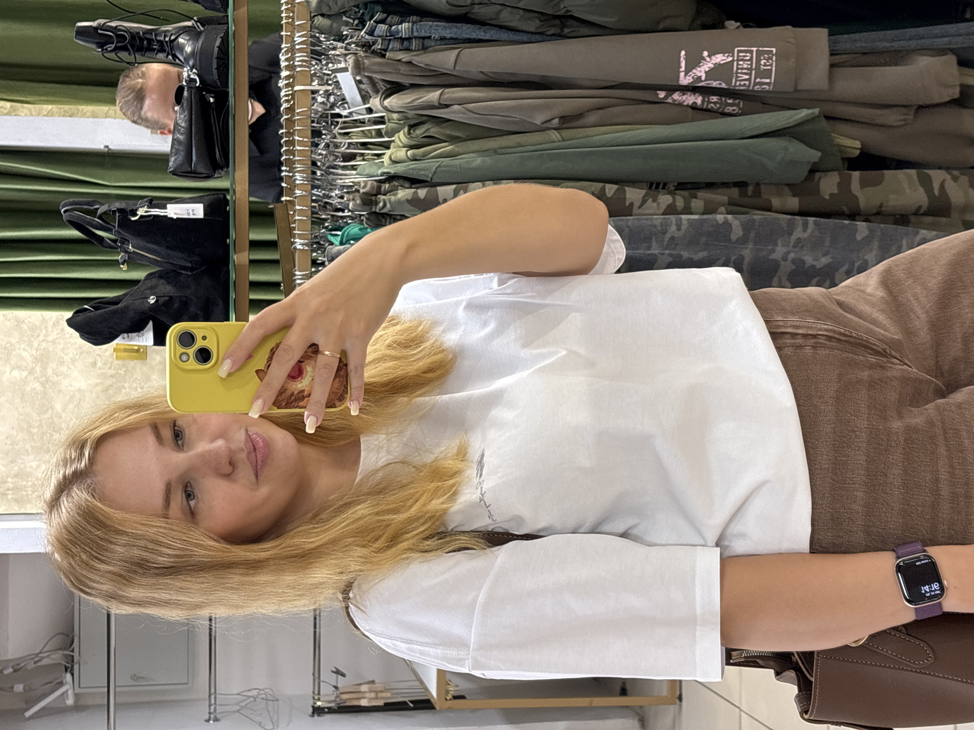 In what appears to be a retail environment, a young woman with blonde hair is taking a selfie in a mirror. She is wearing a white t-shirt, brown trousers, and a dark smartwatch on her left wrist. She is holding a yellow smartphone in her right hand. In the background, a clothing rack filled with various garments, including camouflage-patterned items and green shirts, is visible. Several handbags and a pair of black boots are displayed on a shelf behind the clothing rack. Another person, partially obscured, can be seen in the reflection of the mirror behind the woman. The overall setting suggests a clothing store or boutique, potentially located in Rusanovo, Russia.