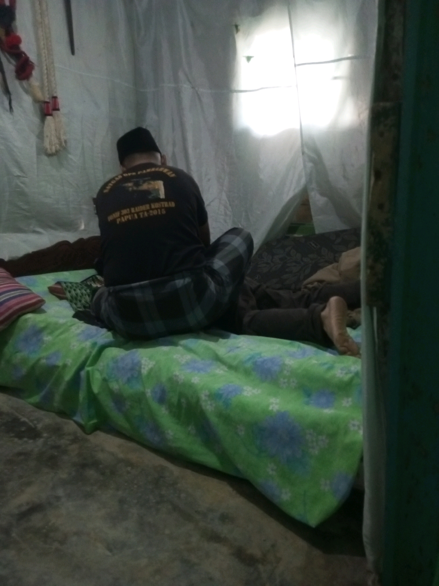 In a dimly lit interior, a person wearing a black skullcap and a dark t-shirt with text on the back is seated on a mattress. The t-shirt has an image and text that reads "BUNU 303 RAIDER KOTRAD PAPUA TA-2018". The person is wearing plaid pants. The mattress is covered with a green sheet featuring a blue and white floral pattern. A pillow with horizontal stripes of pink, white, and blue is visible to the left of the mattress. Another person, partially obscured, is lying down on the mattress with their legs and feet visible. The scene appears to be within a makeshift shelter, possibly a tent, with white sheeting forming the walls. A brightly lit window or light source is visible through the sheeting in the background. The floor is a rough, concrete-like surface. The image was taken in Andongsili, Indonesia.