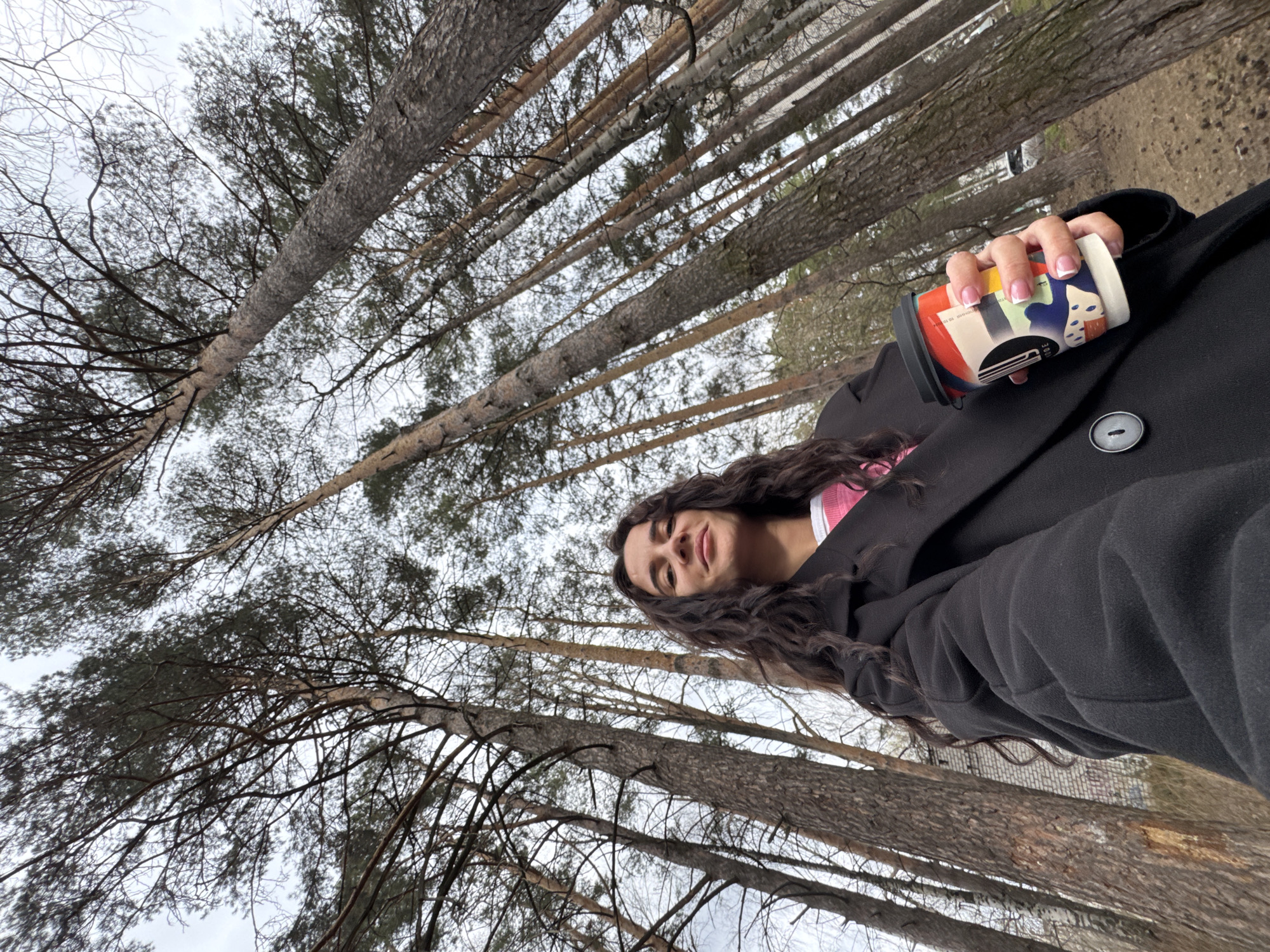 The image depicts a woman in a black coat, holding a patterned cup in her right hand, standing outdoors in what appears to be a park or wooded area. The camera angle is from below, looking up at the woman and the surrounding trees. The trees are tall and bare, with many thin branches visible against a cloudy sky. The cup in her hand has a colorful design with geometric shapes and possible abstract figures. The woman has long, dark curly hair and is looking towards the camera with a slight smile. The background shows a mix of trees and sky, suggesting an outdoor location. The image does not offer enough information to confirm the location is Chaikovsky, Russia, or to make any further forensic deductions.