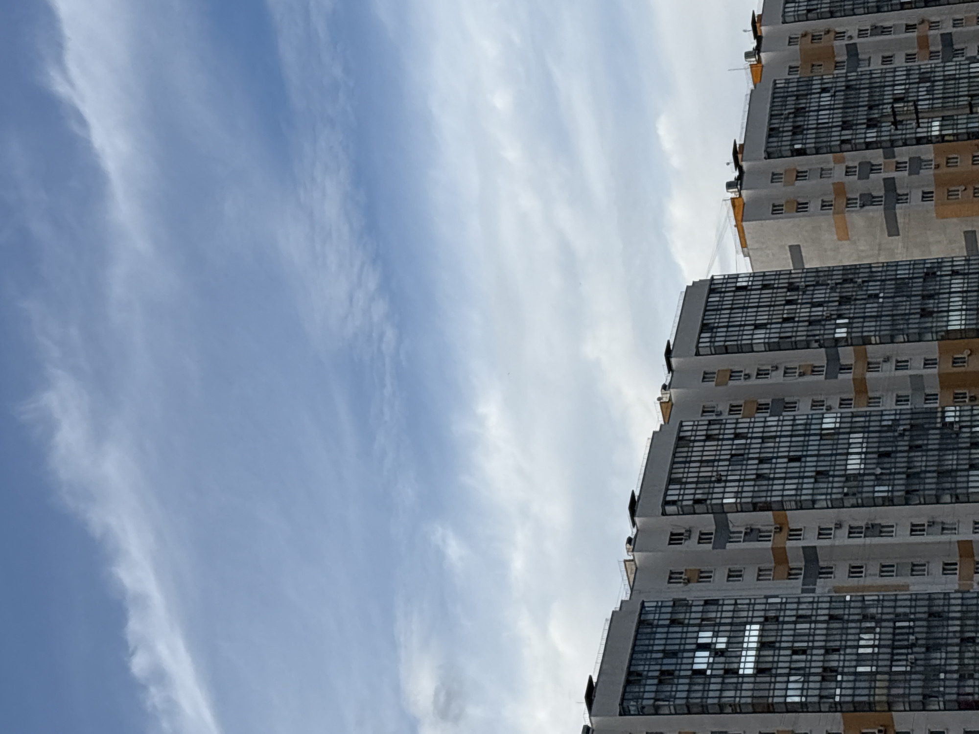 The image displays a close-up, low-angle view of a modern apartment building against a cloudy sky. The building is tall and appears to be constructed with concrete and glass. Its facade is a mix of light gray concrete sections and darker, grid-patterned glass windows, suggesting a contemporary architectural style. There are also horizontal bands of a light orange or yellow color accentuating certain sections of the building. Small, rectangular windows are visible interspersed between the larger glass sections on each floor, and some of these have air conditioning units installed beneath them. The building is segmented into distinct levels, with visible rooflines and parapets. The sky is partly cloudy, with streaks of white clouds against a pale blue background, indicating daylight. The orientation of the photo, with the building dominating the right side and the sky the left, suggests a dynamic, upward perspective. There is no visible text within the image. The scene evokes a sense of urban life and modern construction. Based on the location context, this is likely an apartment complex in Tarasovka, Russia. The weather appears to be overcast or partly cloudy, and the time of day is daytime.