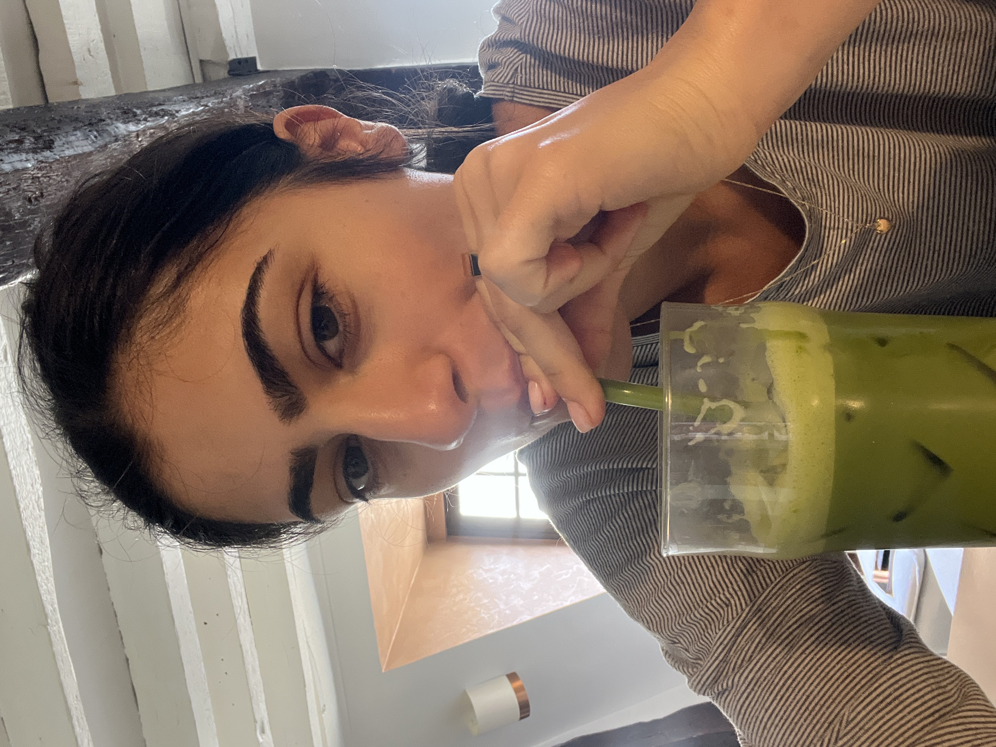 A selfie shows a woman holding a tall glass of a green drink, likely a matcha latte, with a straw. She is resting her cheek on her hand. The woman has dark hair pulled back, dark eyebrows, and is wearing a striped top with a gold necklace. The background includes architectural details indicative of an older building: white, parallel beams on the ceiling and a window with a light-colored frame and a dark cross-member. There is a wall visible with a light brown paint color. A light fixture with a copper band is also present on the wall. While definitive identification is impossible, architectural details suggest a Parisian location.