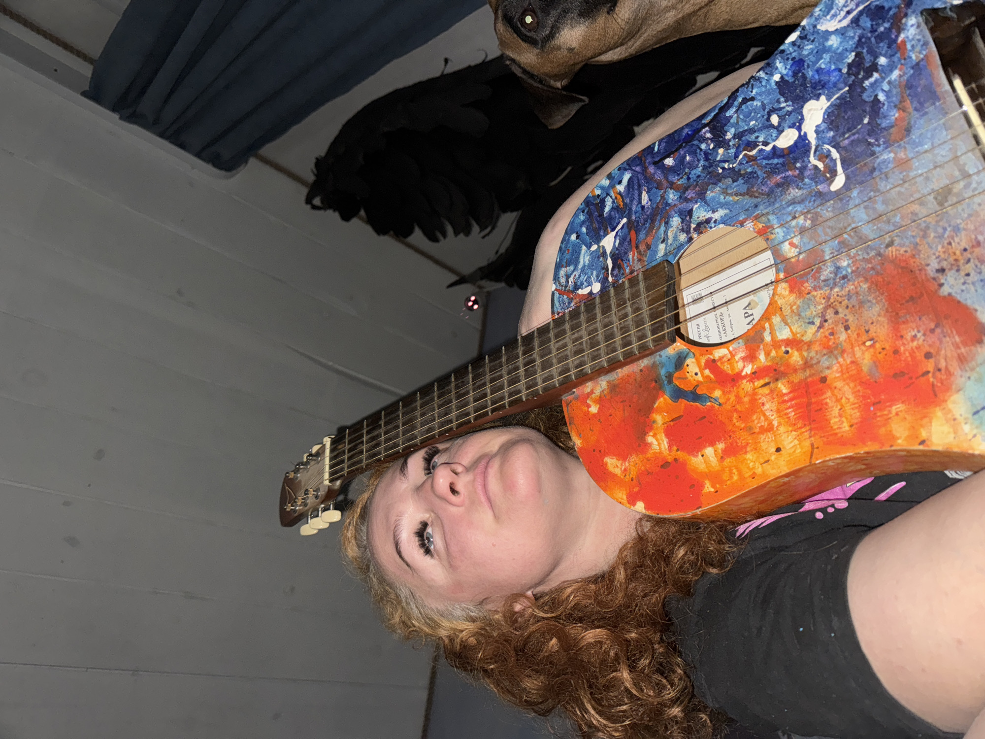 A low-angle shot shows a woman with curly red hair lying down, holding a brightly painted acoustic guitar. The guitar body is primarily orange and blue with white splashes, and the neck stretches upwards, nearly bisecting the frame. A dog's head is visible in the upper right corner, partially obscured. A black object, possibly a bag or folded fabric, sits behind the guitar and the woman's head. The background consists of a white wall and a blue curtain or sheet. A label on the guitar shows lettering that includes Russian text: "РОССИЯ...г. Чайковский...улица Ленина, 63/1...Торговая площадь 1". This indicates the guitar's place of origin as Chaikovsky, Russia.