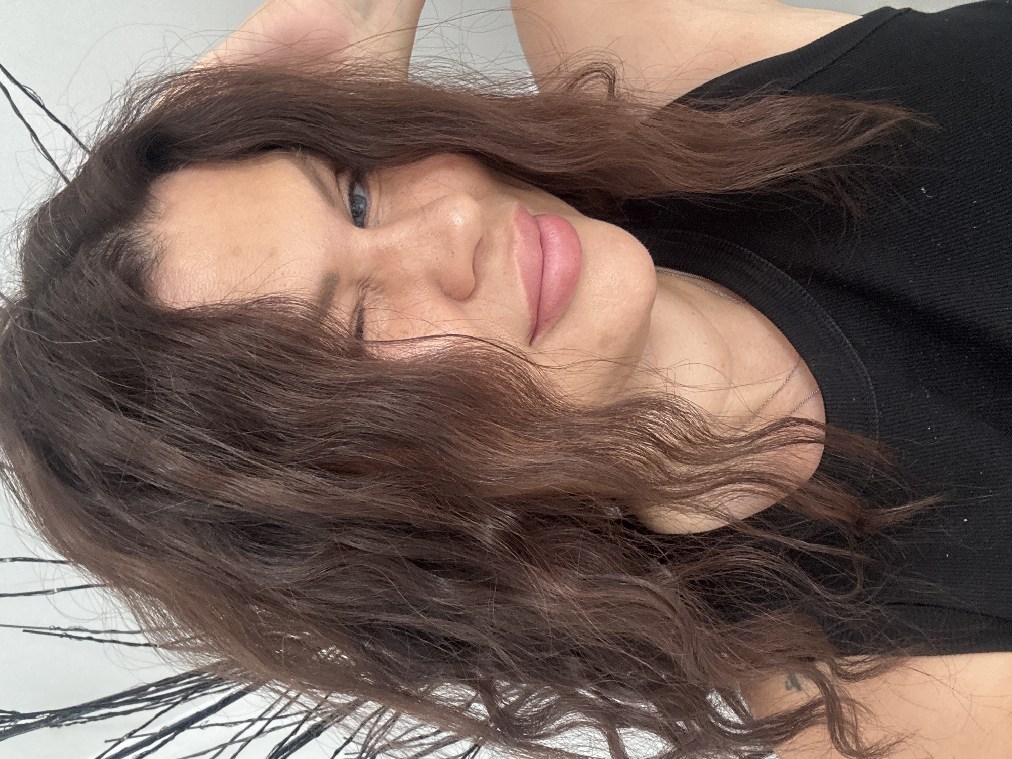 The image is a close-up, waist-up shot of a woman indoors, possibly in a home. She has medium-length, wavy brown hair and blue eyes, and is winking at the camera. Her lips are slightly parted and appear to have pink lipstick. She is wearing a black tank top and a silver necklace. The background is a plain, light-colored wall with some dark, thin branches or twigs visible to the left of her head. There is no indication of the specific location within Katowice, Poland.
