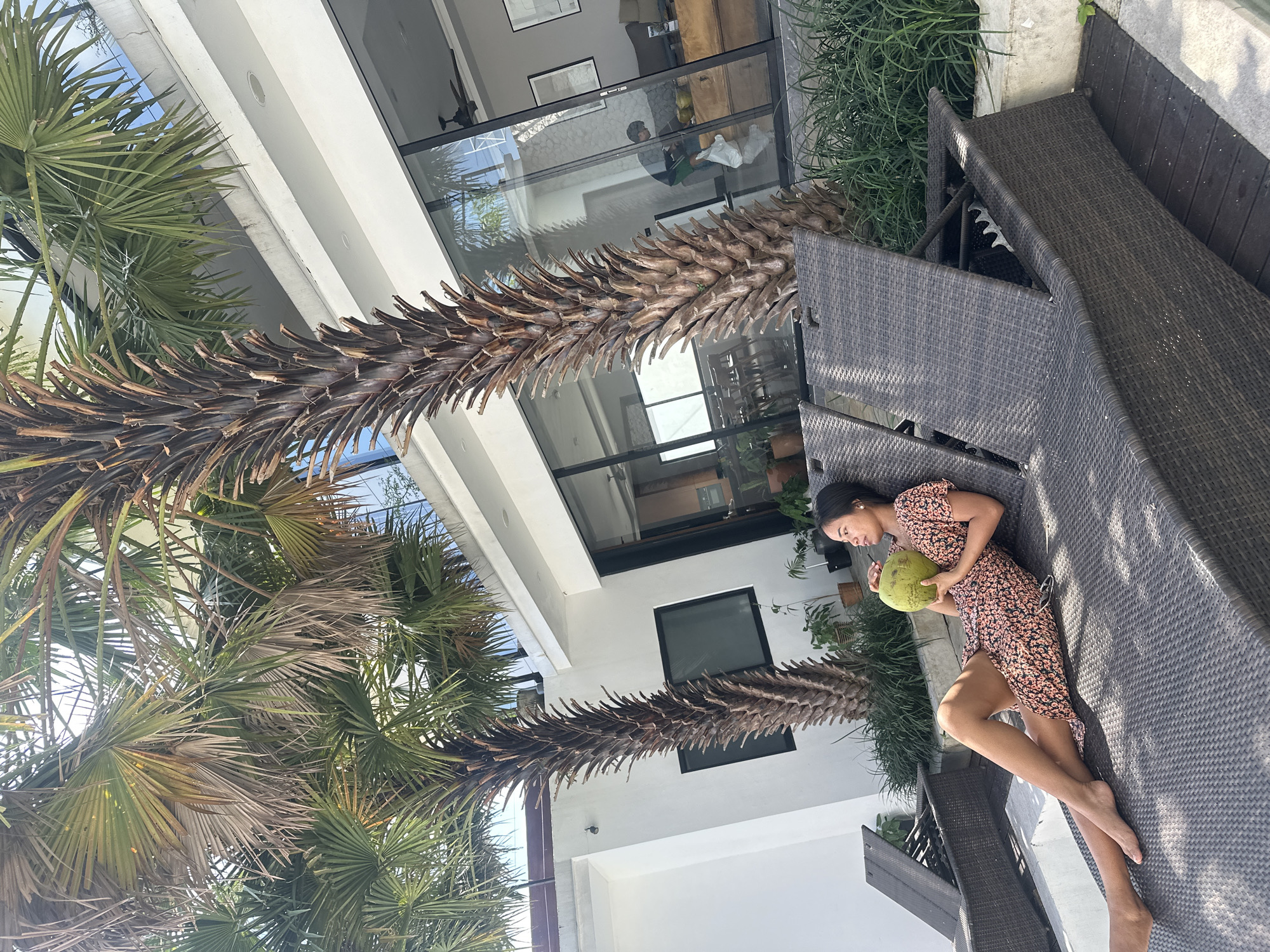 A woman lies on a lounge chair, holding a coconut. She is wearing a floral dress and appears relaxed. The lounge chair is made of woven material and is situated outdoors, likely in a tropical setting given the presence of palm trees. In the background, there is a modern building with large glass windows and doors, suggesting it could be a resort or a private residence. The environment is sunny, casting shadows from the palm trees. The location is identified as North Kuta, Indonesia, which is consistent with the visual cues of palm trees and a warm climate.