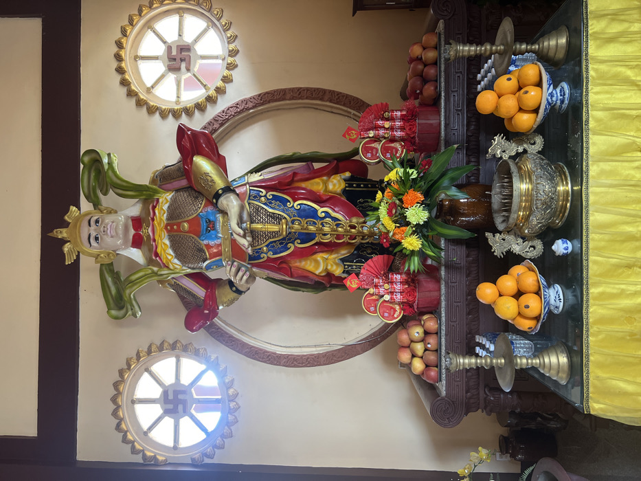 An indoor scene in Đăk Mar, Vietnam, features a large, colorful statue of a standing warrior figure or deity. The statue occupies the central upper portion of the image, depicting a figure with light skin, a golden winged crown, and elaborate armor in shades of red, gold, blue, and green, holding a sword vertically with both hands. Behind the statue is a large, brown, circular architectural element. On either side of the statue, two circular windows are visible, each featuring a swastika symbol (Buddhist symbol) within its frame and surrounded by a gold, petal-like border.
Below the statue, a two-tiered altar or display table is arranged. The upper tier, a dark carved wooden surface, presents offerings including a central brown vase holding a vibrant floral arrangement of orange, yellow, and green flowers with green foliage. Flanking the flowers are two decorative arrangements constructed from stacked red Coca-Cola cans, adorned with red festive elements and small red fans. Piles of red apples are placed on the far left and right ends of this tier.
The lower tier consists of a glass-top table draped with a pleated yellow cloth featuring a decorative border. On this tier, a large, ornate golden incense burner with dragon-shaped handles is centrally positioned. To its left, a pile of oranges rests in a blue and white decorative bowl next to a tall, multi-tiered brass candelabra/stand and a small blue and white ceramic container. To the right of the incense burner, another pile of oranges is in a matching blue and white bowl, with a second brass candelabra/stand partially visible on the far right and a row of small white bottles (possibly water) behind the oranges. The background walls are a plain, light beige color.