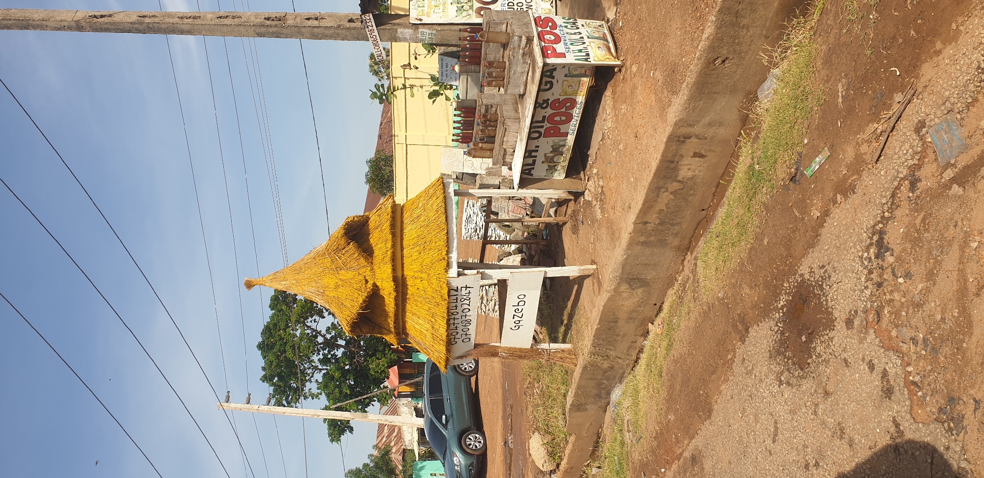 The image depicts an outdoor scene in Barnawa, Nigeria, characterized by a small, thatched-roof gazebo structure. The gazebo is made of what appears to be dried grass or straw, giving it a rustic and traditional appearance. Attached to the front of the gazebo is a white sign with handwritten text that includes phone numbers and the word "Gazebo." To the right of the gazebo, there is a table displaying numerous small bottles filled with a liquid, likely oil or fuel, as well as a POS (Point of Sale) machine and a sign that reads "ALH. OIL & GAS SERVICES." In the background, a green vehicle is parked on a dirt road, and various buildings with corrugated metal roofs are visible. A concrete pole with electrical wires runs vertically through the scene. The ground is a mixture of dirt, gravel, and some sparse vegetation growing along a concrete drainage ditch. The sky is clear and blue, suggesting it is daytime.