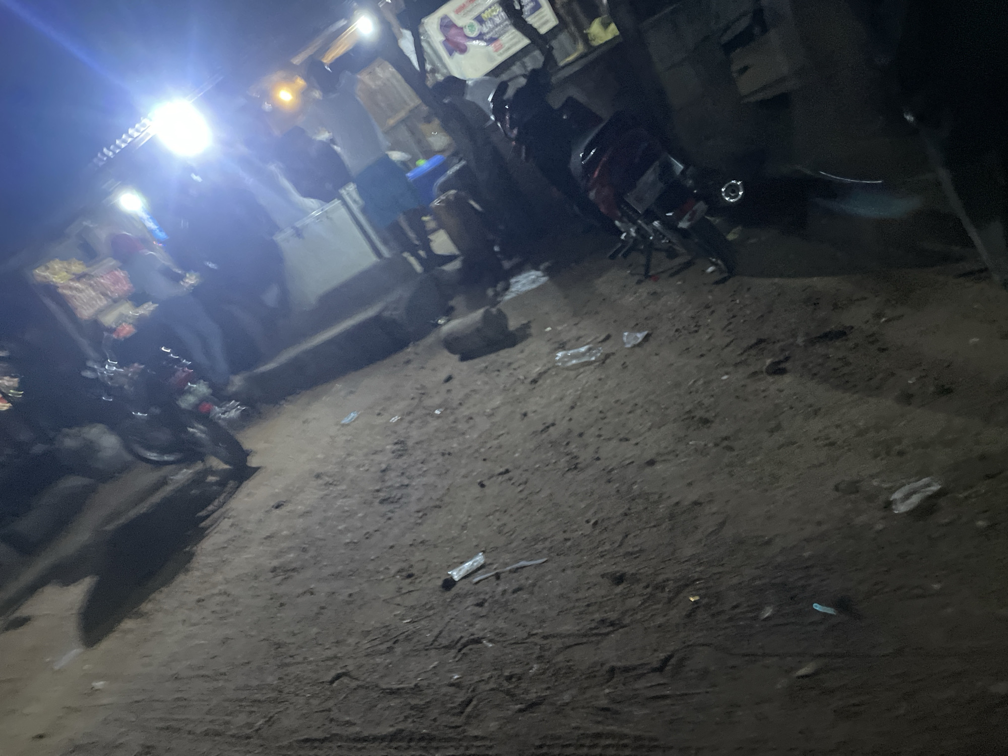 The image depicts a dimly lit, outdoor scene that appears to be a street or roadside area at night, possibly in Lokoja, Nigeria, given the context. The ground is composed of dirt and debris, with some tire tracks visible. Scattered on the ground are various discarded items, including plastic wrappers, bottles, and what looks like a piece of a syringe.
In the background, there are indications of commercial activity or street vending. A bright light source, possibly a street lamp or a strong bulb, illuminates a section of the scene, creating glare and obscuring details. There are also other, less intense lights visible.
Several objects that appear to be motorcycles are present, some parked and others possibly in motion or being worked on. Some figures, likely people, are partially visible, but their features and actions are indistinct due to the low light and blurriness of the image. The overall impression is of a gritty, nighttime urban environment with elements of economic activity and potential signs of drug use.