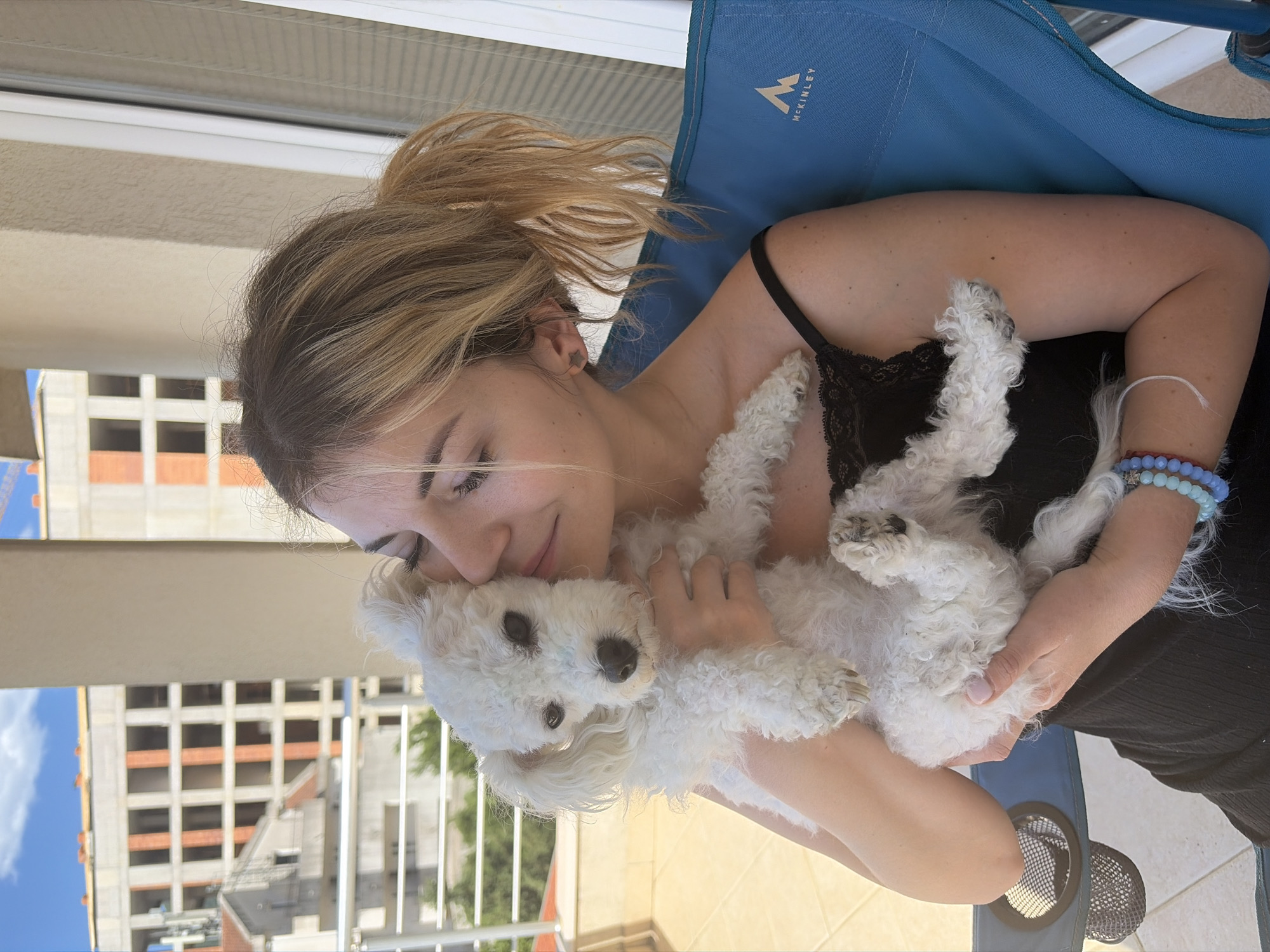 The image shows a light-skinned woman with blonde hair pulled back in a ponytail, holding a small, white dog. She's wearing a black top with lace detailing and sits in a blue McKinley-branded chair. A bracelet adorns her wrist. The background reveals a building under construction with scaffolding, indicative of an urban setting. The scene is bathed in daylight. The presence of a balcony suggests the location is an apartment or similar structure. The architectural style and setting suggest the photograph was taken in Budva, Montenegro. The woman is cuddling the dog against her face.