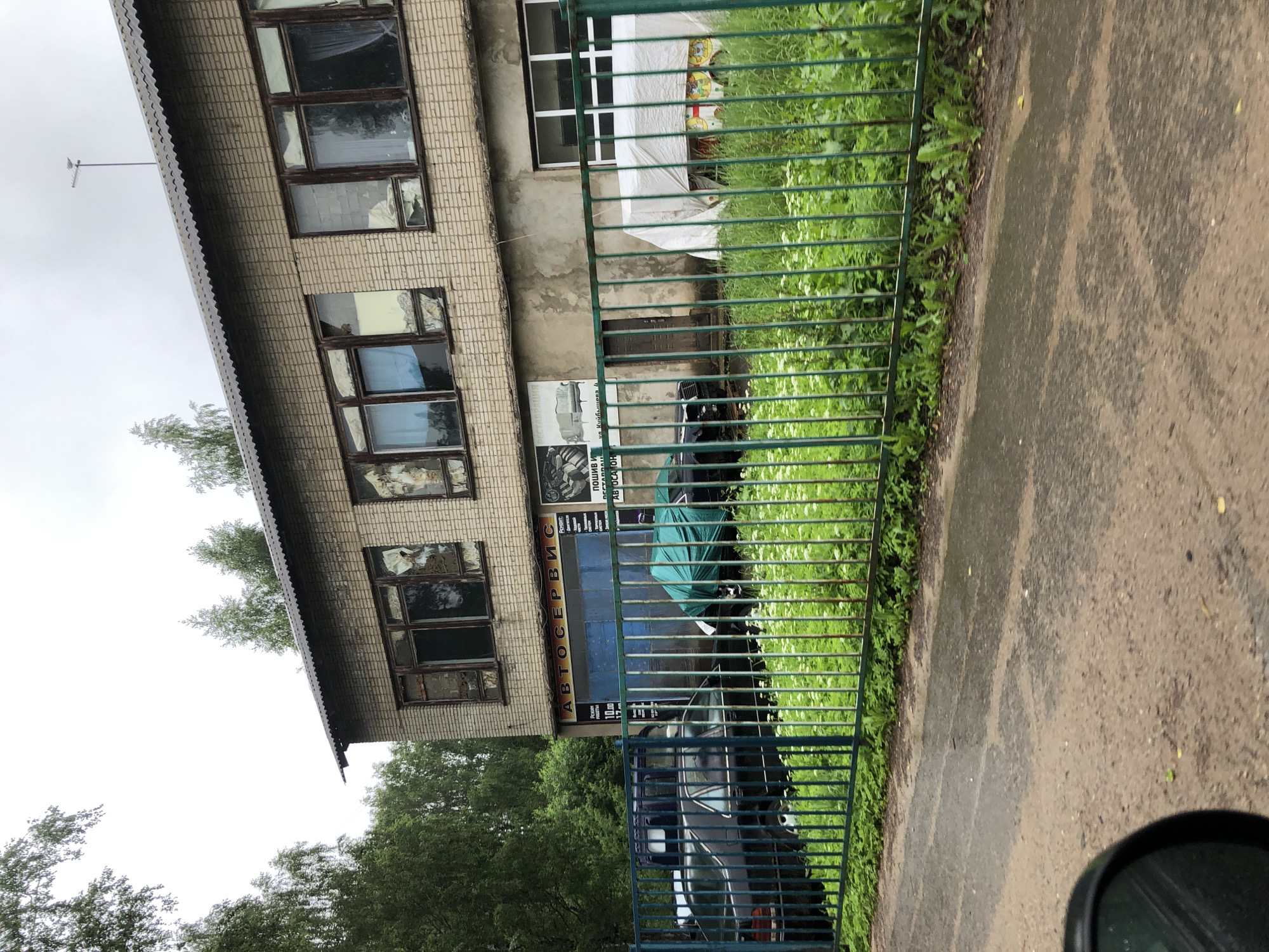An overcast, daytime exterior shot shows a two-story, light-colored brick building with a corrugated metal roof, located in Safonovo, Russia. The building appears to be an auto service center, with large blue garage doors and signage visible on the facade. In front of the building, a green metal fence encloses a grassy area with several parked cars. One car is covered with a green tarp, and another is partially obscured by a white tarpaulin. The ground in front of the fence is a wet, asphalt road, suggesting recent rain. Trees surround the property, and the sky is a uniform grey.