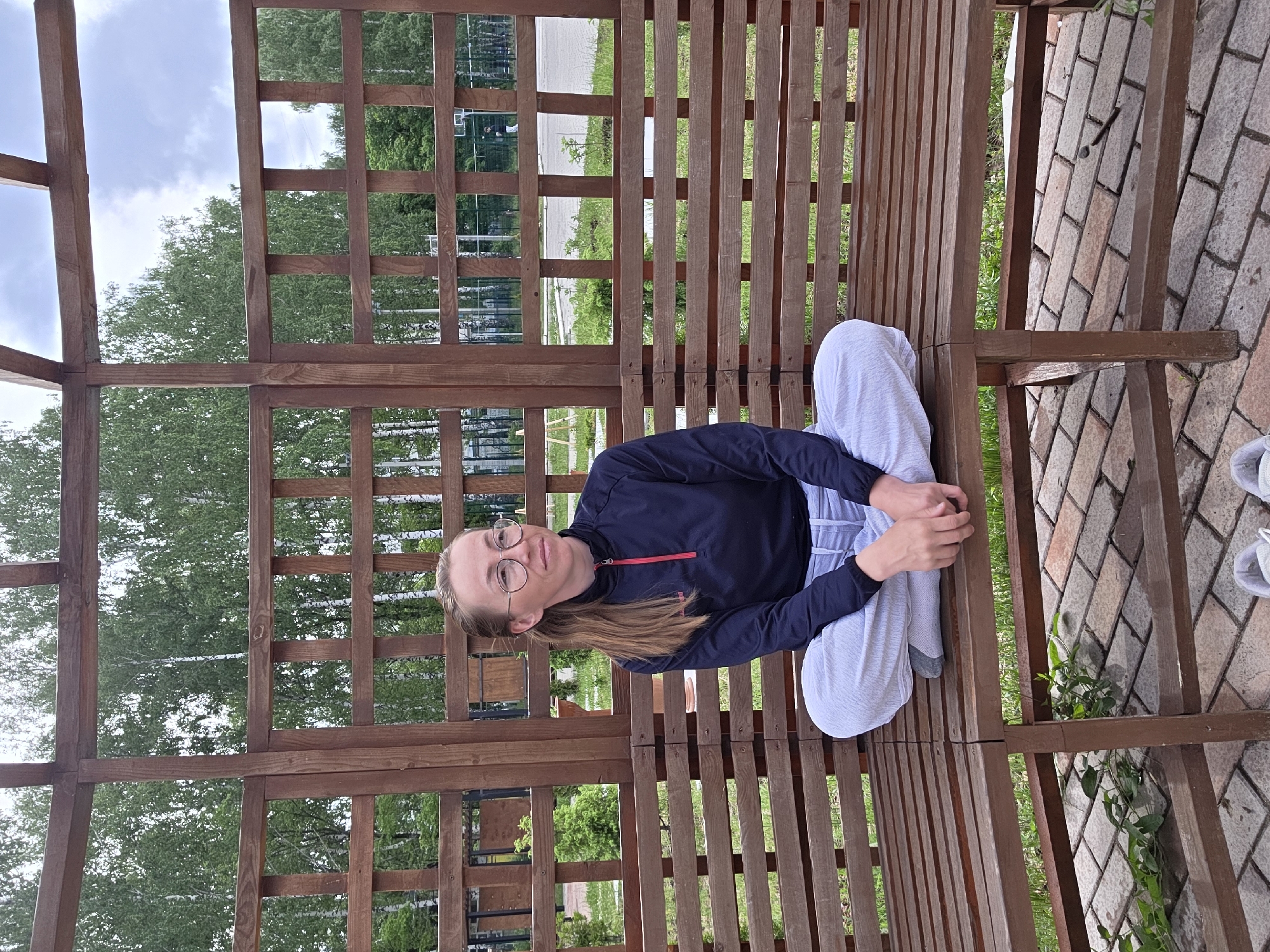 A woman is seated cross-legged on a wooden bench inside a wooden structure with a latticed design. She is wearing glasses, a dark-colored jacket with a red zipper detail, and light-colored striped pants. The bench is located on a brick-paved area with some greenery visible. Trees are visible through the latticed structure in the background, along with a glimpse of a street or pathway. The location is the city of Unknown, Unknown.