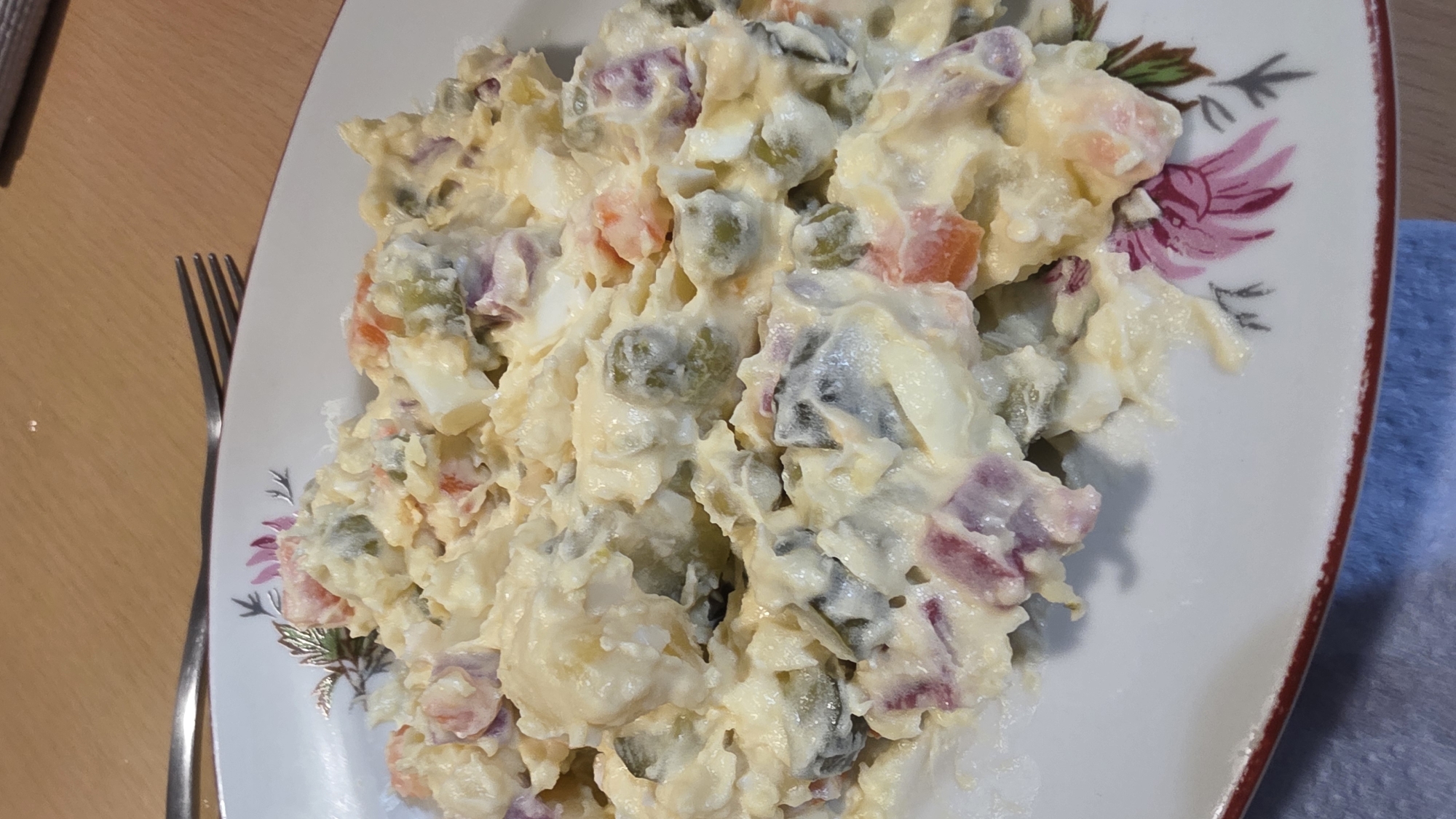The image presents a close-up, overhead view of a plate of potato salad, positioned on a light-brown wooden table. The main subject is a creamy, mayonnaise-based potato salad generously served on a white plate with a decorative floral pattern.
The salad appears to be composed of various diced ingredients, including soft chunks of light-yellow potatoes, white pieces of hard-boiled eggs, small round green peas, and orange pieces of diced carrots. There are also darker, purplish-red fragments mixed throughout, which could be diced red onion or pickled vegetables. The dressing evenly coats all the ingredients, giving the salad a rich and textured appearance.
The white plate features a distinct floral design along its rim, with stylized flowers and leaves in shades of pink, green, and brown. A thin, reddish-brown line borders the outer edge of the plate. To the left of the plate, part of a silver-colored fork is visible on the wooden table, implying the meal is ready for consumption. To the right of the plate, a blurred section of a blue cloth or napkin can be seen.
The scene suggests an indoor setting, likely a home kitchen or dining area. No people are visible, and there is no text present in the image. The time of day and weather cannot be determined from the visual information provided. While the external context indicates Blagoevgrad, Bulgaria, there are no specific visual cues within the image itself to confirm this location.