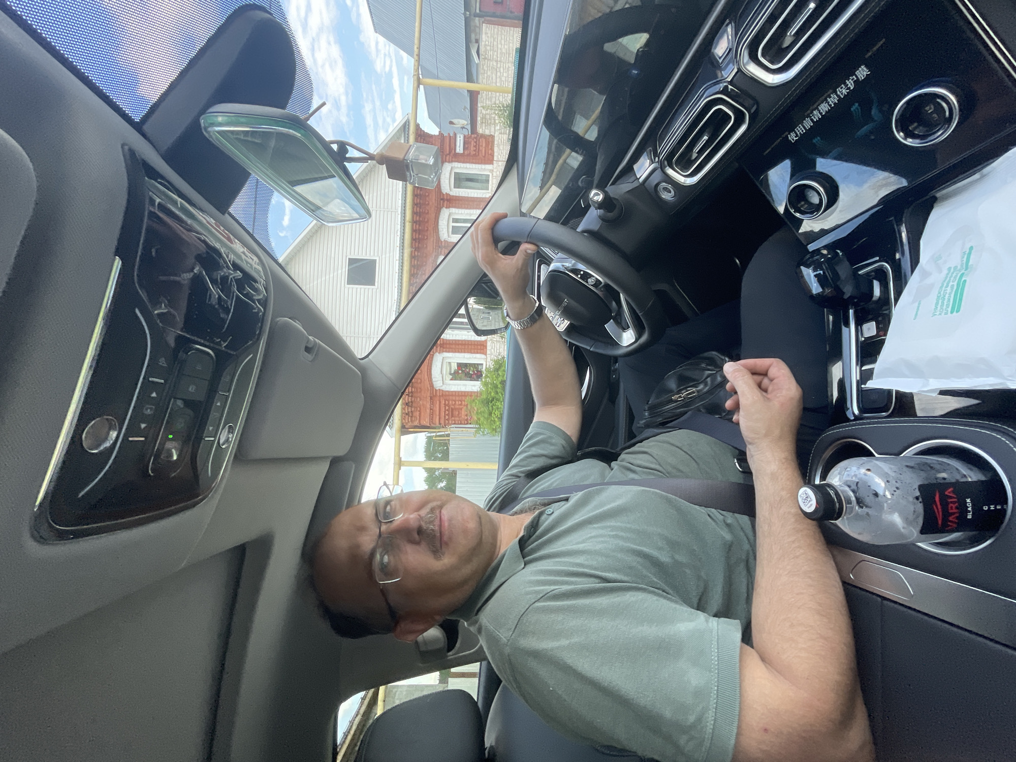 A middle-aged man with a mustache and glasses is seated in the driver's seat of a car, looking towards the camera. He is wearing a green polo shirt and a seatbelt. His right hand is on the steering wheel. The car's interior is visible, including the dashboard, rearview mirror, and a cup holder with a bottle of water. Outside the car, a rural scene with houses and a blue sky can be seen. The location is identified as Maminskoye, Russia.