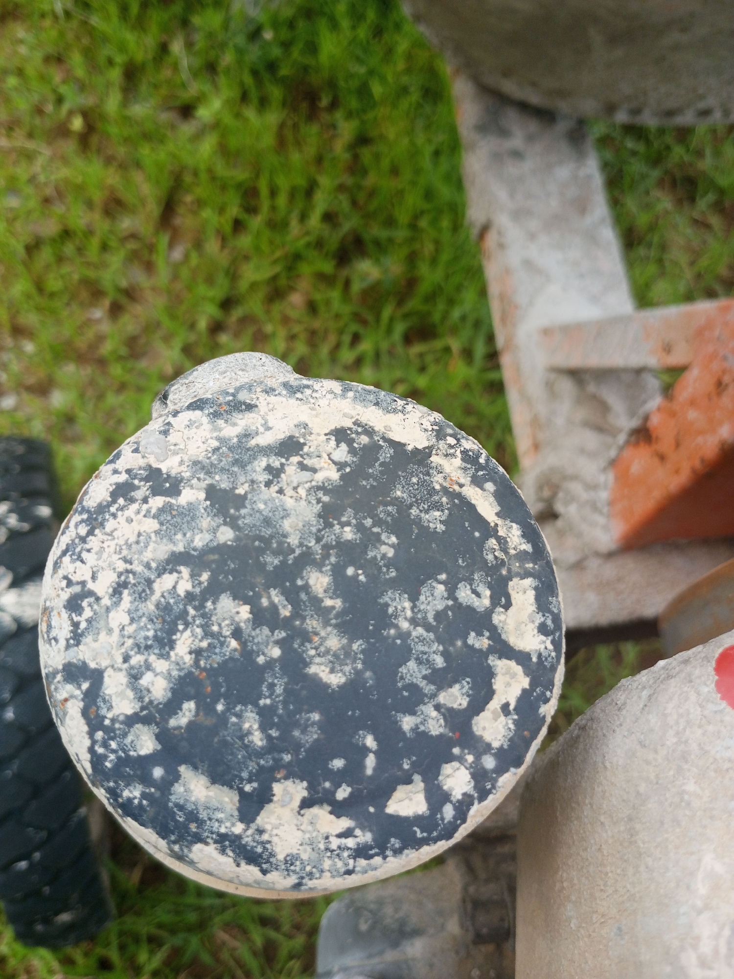 The image shows a close-up of a large, round, dark object with a rough, textured surface. This object is speckled with white and light gray material, suggesting it is coated or splattered with something like cement or paint. It appears to be part of a larger structure, possibly construction equipment, given the visible parts of what looks like a tire and other metal components in the background. The setting is outdoors, with green grass and soil visible in the background, indicating it is daytime. The lighting suggests an overcast or cloudy day. The focus is on the detailed texture of the round object, which shows signs of wear and use. There are no people or visible text in the image. The overall impression is of a piece of industrial or construction machinery in a rural or outdoor environment.