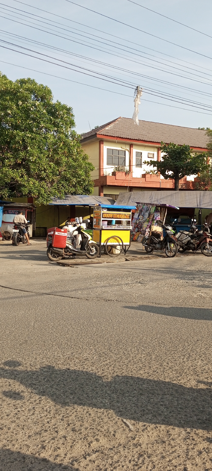 The image depicts a street scene in Setu, Indonesia, likely during the daytime, with a building in the background and a road in the foreground. The building appears to be a commercial or institutional structure with a light yellow facade, red trim, and a dark tiled roof. It has multiple windows, some with vertical blinds. In front of the building, there are several makeshift stalls or carts, one of which is yellow and features signage that includes "SUSU KEDELAI". There are also motorcycles parked and in use. One motorcycle in the foreground is white with a red cargo box labeled "Yakult". Another motorcycle is parked in front of the yellow cart. A person is seated on the white motorcycle. To the left, another person is riding a motorcycle away from the camera. The road surface is asphalt with a textured pattern. Overhead, power lines are visible against a pale sky. Trees with green foliage and white flowers are also present, particularly a large one to the left. Shadows are cast on the road, suggesting the sun is at an angle. The overall impression is a busy street with vendors and transportation.