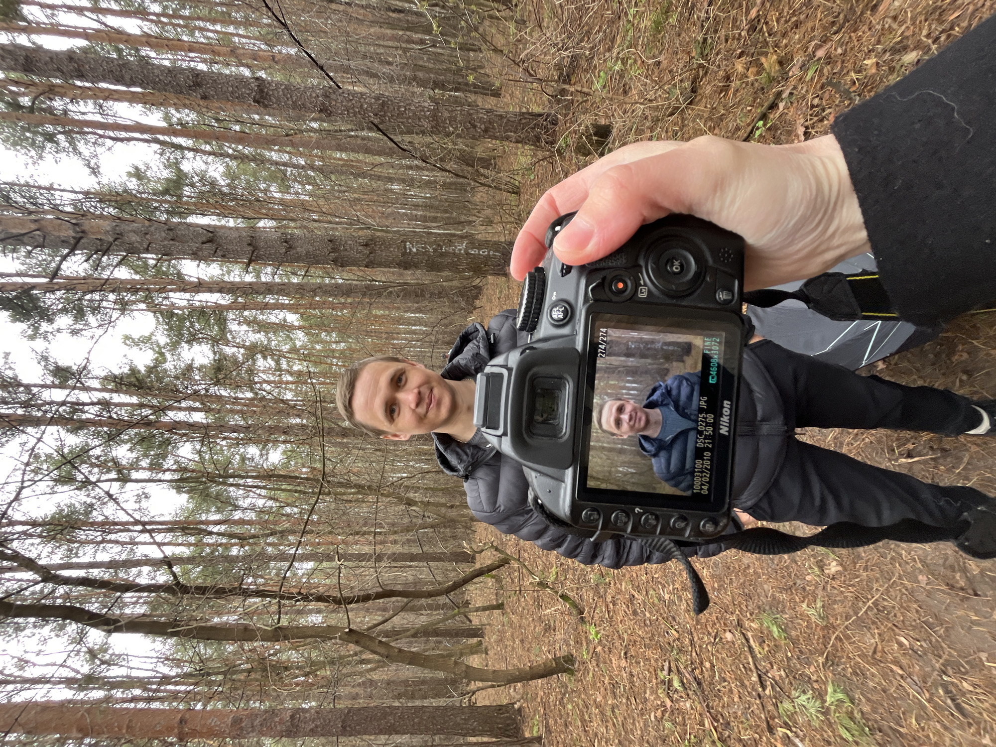 The image, displayed rotated 90 degrees clockwise from its natural orientation, captures a moment of photography in a forest setting, likely in Chaikovsky, Russia. In the foreground, a person's hand, partially covered by a black sleeve, holds a black Nikon DSLR camera.
The camera's display screen prominently shows a photograph of a man. This same man, the apparent subject of the current photoshoot, is also visible standing in front of the camera, looking directly at the viewer with a slight smile. He is dressed in a dark blue puffer jacket and black pants. On the camera screen, several pieces of information are visible: "100D3100 DSC_0275.JPG", "04/02/2010 21:56:00", "FINE", and "4608x3072", along with the "Nikon" logo and "EV". There's also "274/274" in the top right. This data indicates the photo displayed on the screen was taken at 9:56 PM on April 2, 2010, although the ambient lighting of the present scene suggests daytime under an overcast sky.
The background is a dense forest with numerous tall, slender trees, likely pines or similar evergreens, many of which have sparse foliage or bare branches, indicating a late autumn or early spring season. The forest floor is covered with a layer of brown fallen leaves and pine needles. The sky, visible through the tree canopy, is bright and uniformly overcast, providing soft, diffused lighting. A black and silver object, possibly a photographic reflector or stand, is partially visible on the ground near the photographer's feet. Faint white graffiti, seemingly starting with "Nev", can be seen on a tree trunk in the mid-ground.