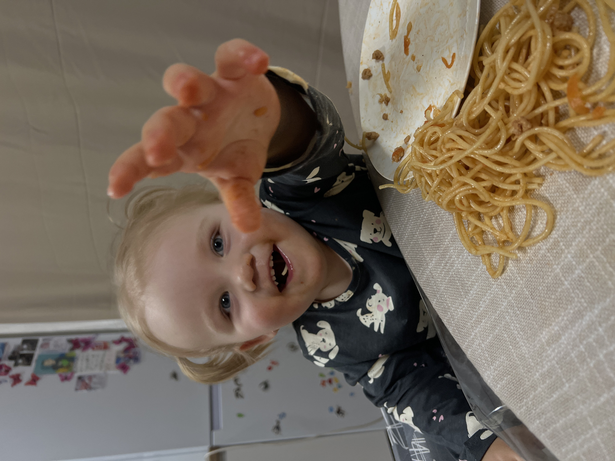 A young child, appearing to be a toddler, is seated at a table in what is identified as a domestic kitchen or dining area in the city of Leninakan, Russia. The child has blonde hair styled in two small pigtails, blue eyes, and a broad smile, with food residue visible around the mouth and a piece of food on the tongue. The child is wearing a dark long-sleeved shirt patterned with white animal figures. The child's right hand (viewer's left) is extended forward, appearing blurred due to motion and covered in orange food residue. On the table, a partial white plate containing remnants of spaghetti with sauce and ground meat is visible, alongside a substantial pile of spaghetti with sauce and ground meat scattered directly on a light brown, patterned tablecloth. In the background, a white refrigerator adorned with magnets, papers, and photos is situated to the left of the child, next to a plain light-colored wall.