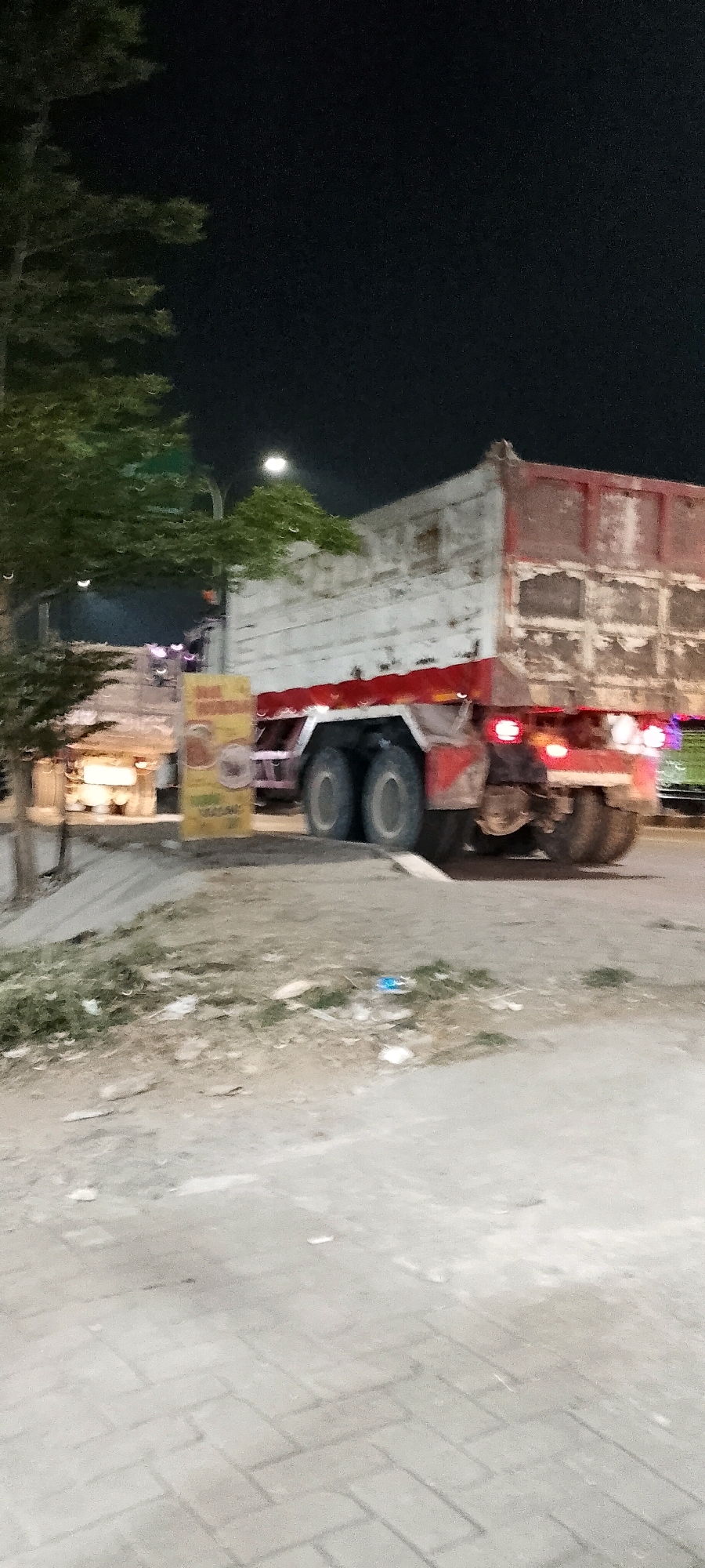 In the city of Cileungsi, Indonesia, at nighttime, a large, weathered dump truck with a white and red body is parked on a paved area next to a road. The truck's rear is visible, showing its large dual wheels and red taillights. To the left of the truck, there is a yellow sign with Indonesian text and images, possibly advertising food or services. A tree with green foliage stands to the left of the sign. The ground in the foreground consists of cracked pavement and loose debris, including some scattered trash. The background is dark with some artificial lights, suggesting an urban or commercial area. The scene appears to be a roadside location, possibly near businesses or a transportation hub.