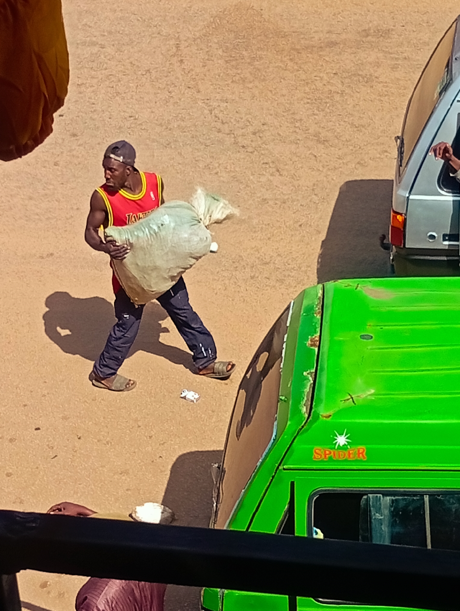 In Bukuru, Nigeria, a Black man wearing a red basketball jersey with yellow trim and a dark baseball cap is carrying a large, light-colored sack. He is walking on a dirt road. To his right, a silver van is parked, and in the foreground, the roof of a bright green vehicle is visible. The ground appears to be unpaved, with scattered debris.