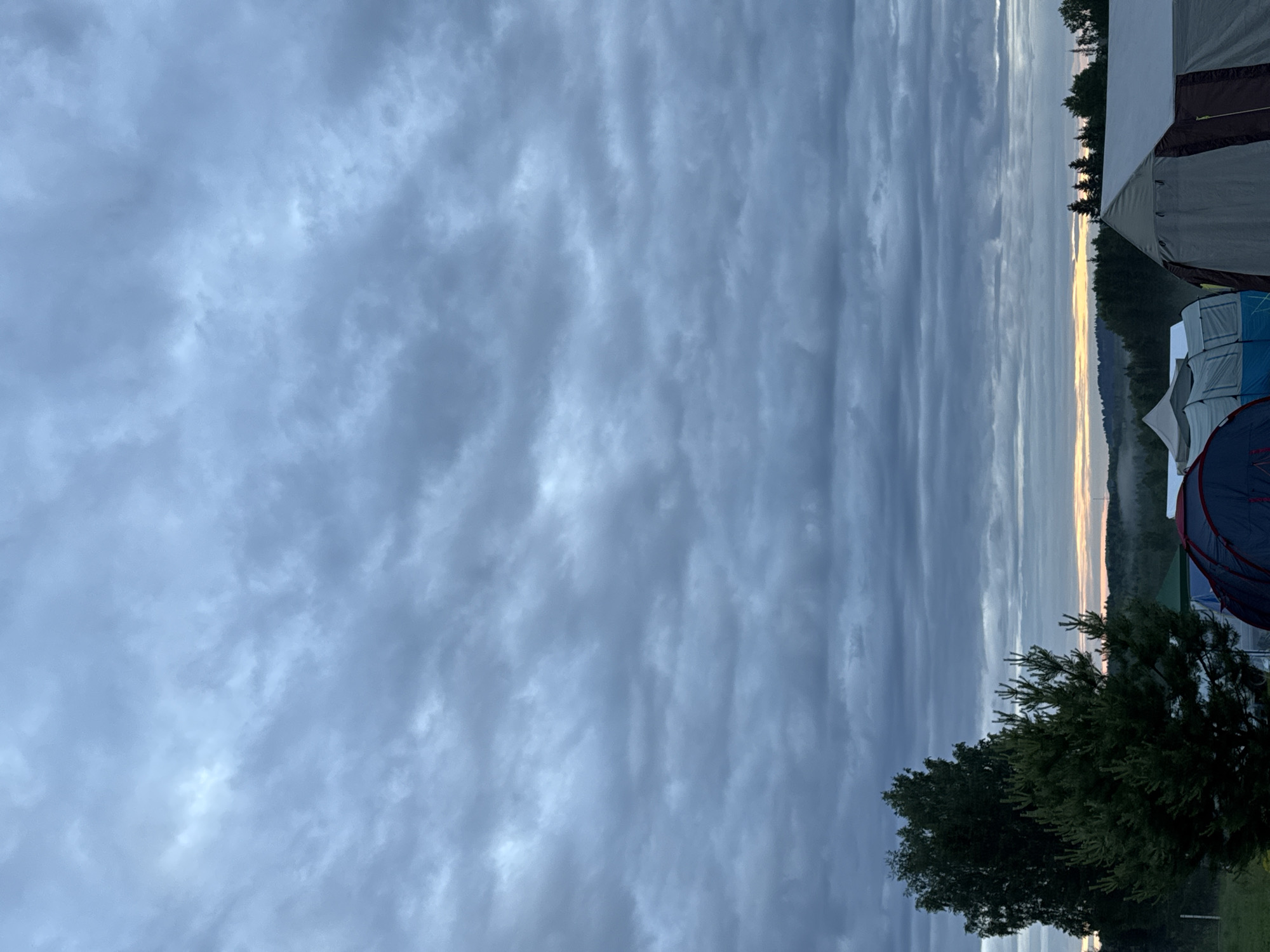 The image depicts a rural campsite near Большая Кивара, Russia, under an overcast sky. The upper two-thirds of the frame are filled with dense, multi-layered grey and muted blue stratocumulus clouds. A narrow band of faint yellowish-orange light is visible on the distant horizon, suggesting either dawn or dusk. The middle ground consists of rolling terrain covered by dense forest, with a light layer of mist partially obscuring trees on the left.
In the foreground, a tall, dark green deciduous tree is on the left, alongside a smaller, bright green coniferous tree. Several camping tents are arranged across the foreground: a dark blue and red tent is partially visible on the far left, light blue and white tents are situated centrally, and a prominent large, multi-section tent in grey and brown occupies the right side. The ground is covered with green grass. No individuals are present in the scene.
