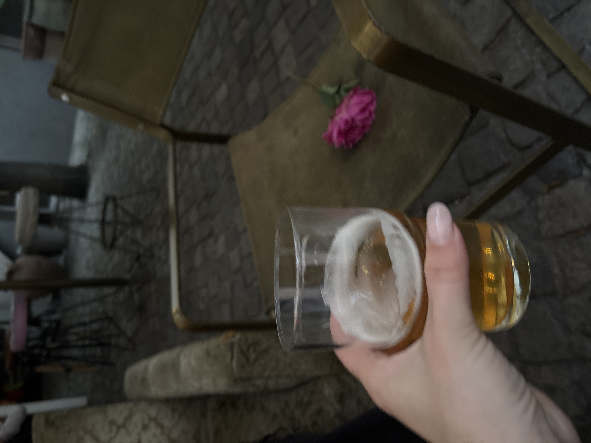 A first-person perspective photograph captures a hand holding a glass of beer in an outdoor or semi-outdoor cafe setting. In the lower left, a fair-skinned hand with light pink manicured fingernails firmly grasps a clear glass filled with an amber liquid topped by a white foam head. In the midground, to the left, a portion of a light-colored, possibly beige or cream, upholstered bench with an intricate pattern is visible. To the right of the beer glass, a chair with a golden-toned metal frame and khaki-colored fabric seat and back is present. A single fuchsia-pink rose, complete with a green stem, lies on the seat of this chair. The ground is paved with dark grey or black cobblestones. The background is out of focus, revealing blurred forms of additional cafe furniture, including stools with dark bases and hints of light or pink seating, and vertical cylindrical elements. The lighting is dim, suggesting an evening or shaded environment. No definitive visual cues in the image identify the location as Sofia, Bulgaria.