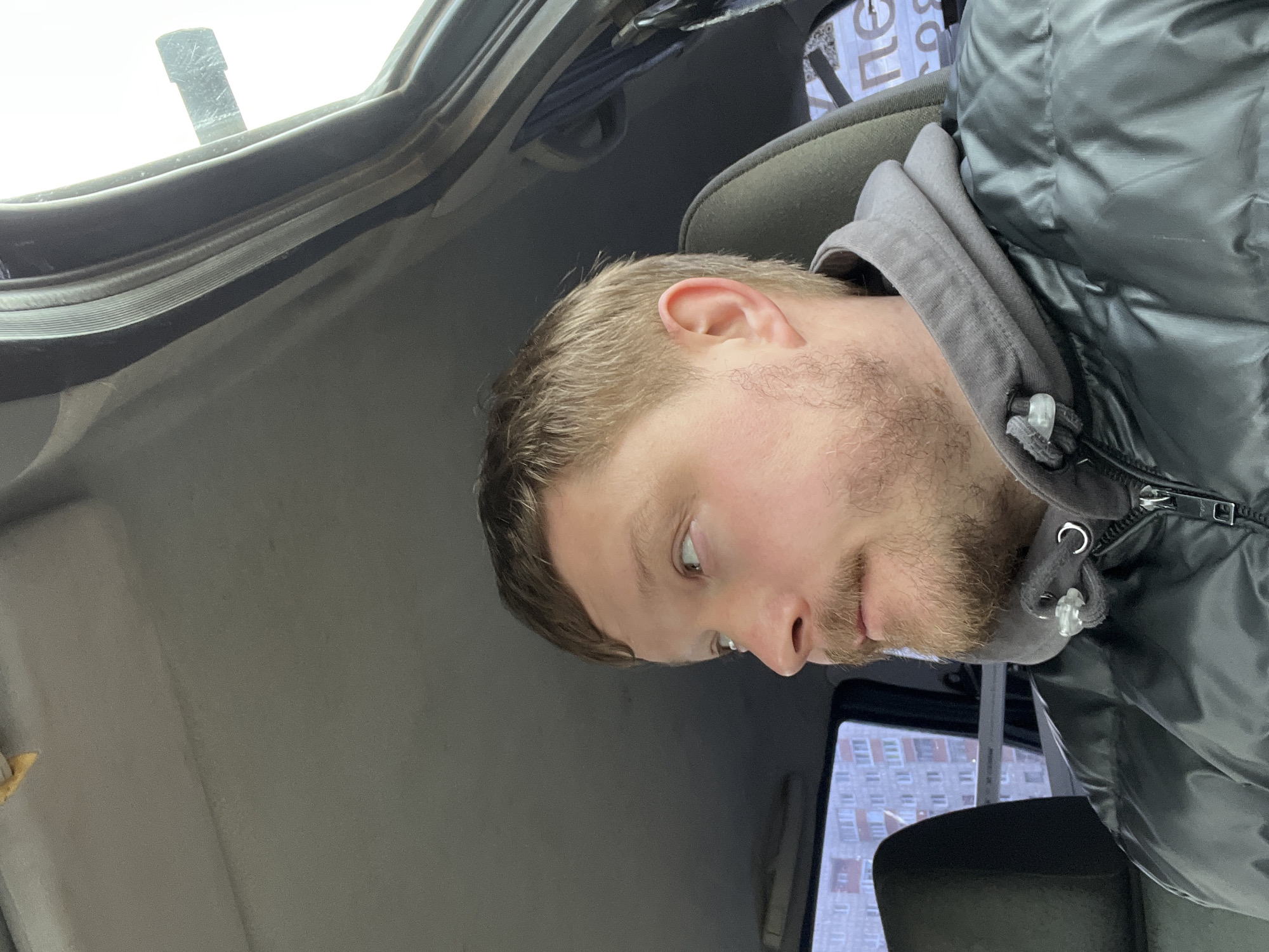 A man with light brown hair and a beard is sitting in a car. He is wearing a gray hoodie and a black puffer jacket. The interior of the car is gray. Outside the car window, a tall residential building is visible. The building is white with rows of windows. The location appears to be Chaikovsky, Russia.