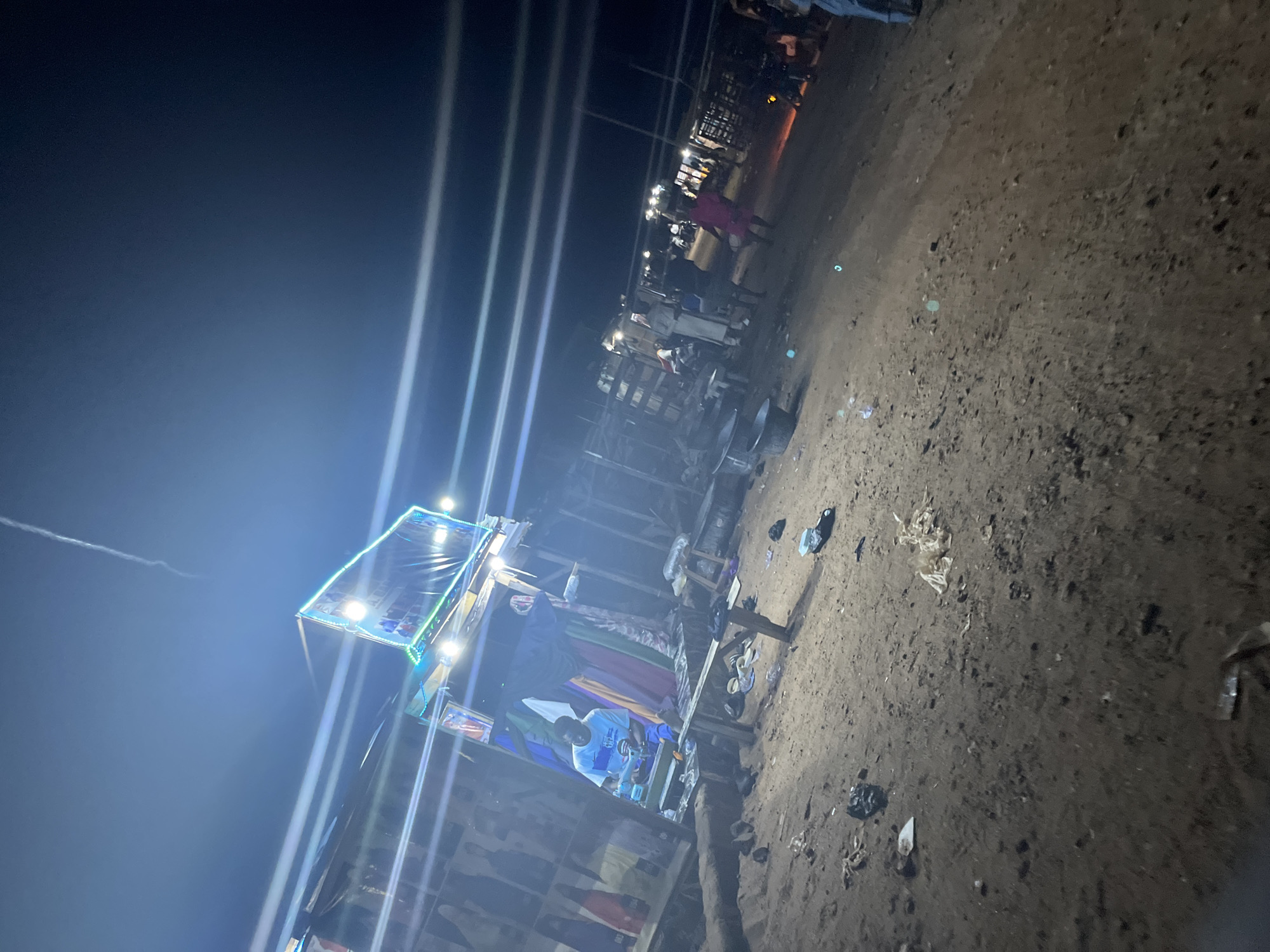 An outdoor, nighttime scene depicts a sandy, dirt ground scattered with debris. In the foreground, a collection of dark, irregular objects, likely rocks or garbage, lies on the ground. To the left, a man wearing a white and blue t-shirt and blue pants sits on a wooden bench. In front of him are various items, including small containers and what appears to be a tabletop. Behind the man, a wooden structure with colorful fabrics draped inside is visible, topped with a bright, illuminated sign featuring text and images. Further into the scene, several people are walking and standing. Some individuals are wearing traditional attire, including a person in a white robe and another in a pink outfit. The background shows more structures and the faint glow of lights, suggesting a market or gathering place. The lighting is harsh and artificial, with prominent light streaks across the image, indicating a camera flash or strong light sources. The overall atmosphere is that of a lively, albeit somewhat disheveled, nighttime outdoor event or market.