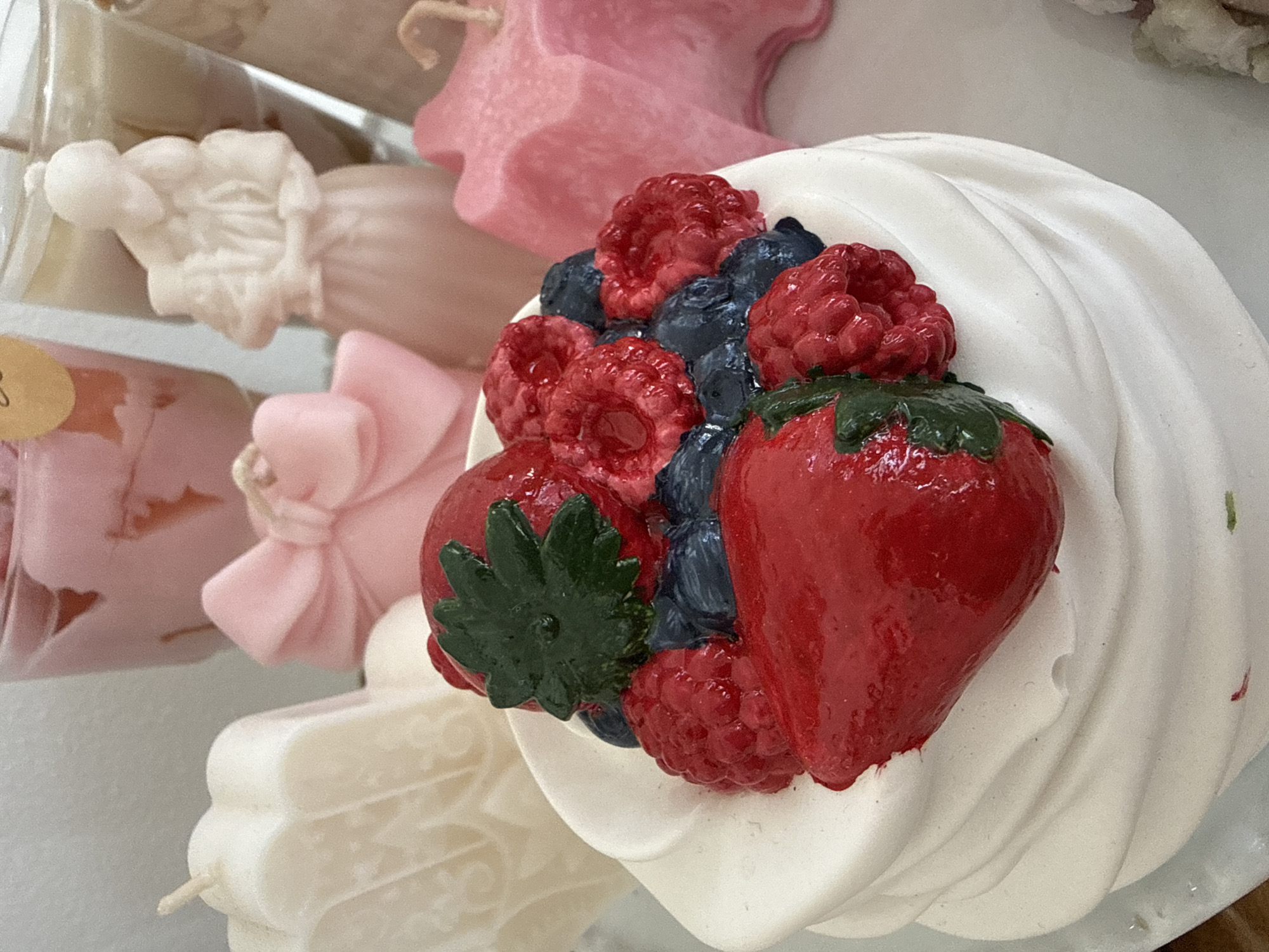 The image displays a close-up of a white, multi-layered candle shaped like a dessert topped with red berries and blueberries. In the background, several other candles are visible, including pink bow-shaped, star-shaped, and a figure of a person holding a baby. The scene appears to be an indoor setting, possibly a shop or a home, with a plain background. There is no indication of a specific location in Tel-Aviv, Israel.