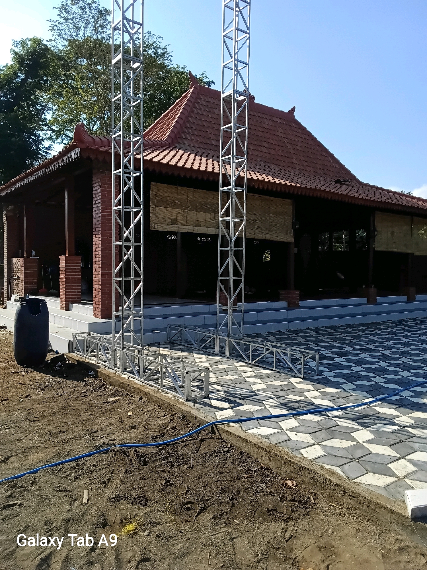 The image depicts an outdoor scene in Trenggalek, Indonesia, possibly a construction site or preparation for an event. In the background, a traditional Indonesian-style building with a reddish-brown tiled roof stands prominently. The building features exposed brick columns and bamboo blinds covering open doorways. In the foreground, two large, metallic truss structures are erected vertically. These structures are made of interconnected metal beams and appear to be part of a stage or support system. In front of the building, a patterned tiled pathway, with alternating light and dark grey diamond shapes, leads towards it. The ground in the immediate foreground is unpaved, with exposed soil and a blue hose running across it. A large, black plastic barrel is visible to the left of the building's entrance. The sky is clear and blue, suggesting fair weather. The overall impression is one of an outdoor area being set up for use, with a blend of traditional architecture and modern construction elements.