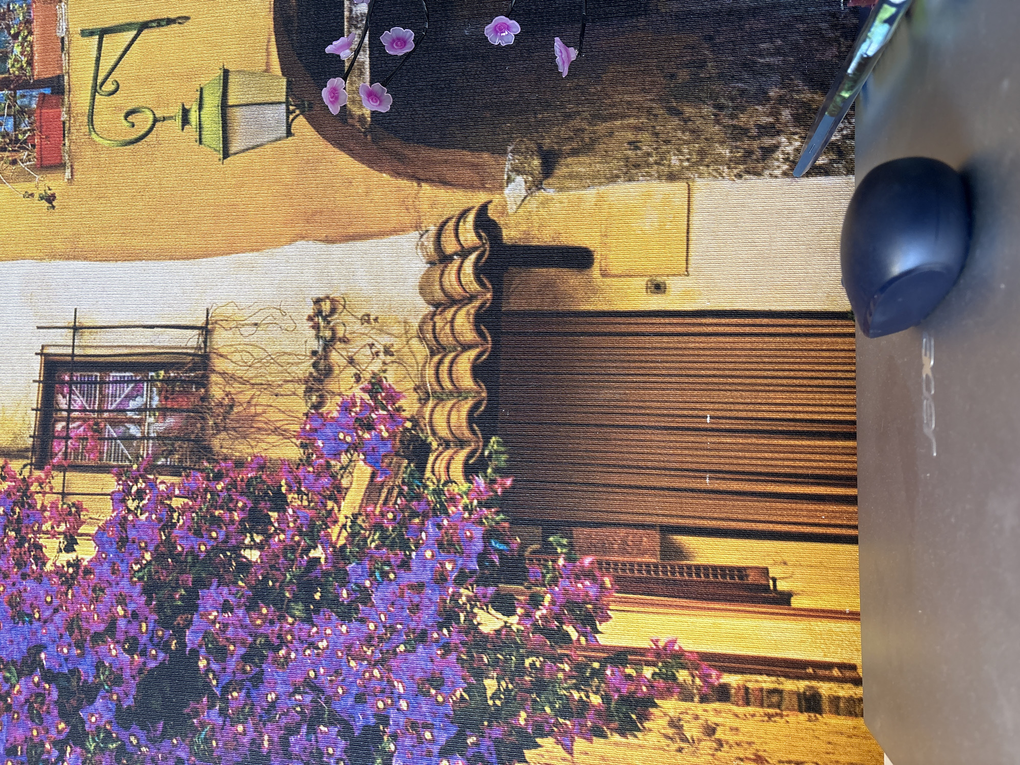 The image displays a close-up view of a printed fabric, likely a tablecloth or tapestry, depicting a vibrant, stylized scene of an old European street. A large bush with abundant purple flowers dominates the left side, cascading down and partially obscuring a brown, paneled door. Above the door is a curved, tiled awning. To the right of the door, a cream-colored wall transitions to an ochre-colored wall. Mounted on the ochre wall is a green, vintage-style lantern with intricate scrollwork. A few small, pink floral accents are visible on the right edge of the fabric.
In the foreground, a dark gray computer mouse rests on a metallic laptop surface. To the right of the mouse, a portion of a pen with a colorful, abstract pattern is visible. The laptop surface has the "acer" logo faintly embossed.
The image does not contain any information to place it in the city of Georgiyevsk, Russia.