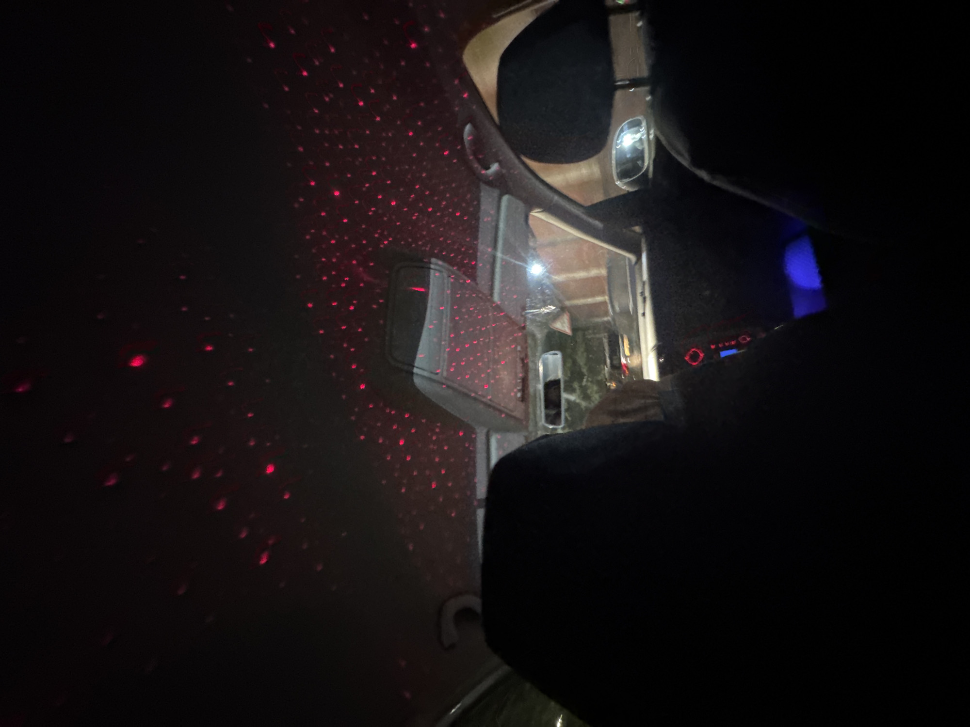 The image shows the interior of a car at night, with red laser lights projected onto the ceiling, creating a starry sky effect. The perspective is from the back seat, looking forward towards the dashboard and windshield. Two dark headrests are visible in the foreground. The dashboard has some illuminated red and blue lights. Outside the car, through the windshield, indistinct shapes suggest a street or parking area with some structures visible. There is no specific information in the image to indicate the location is Chernivtsi, Ukraine.