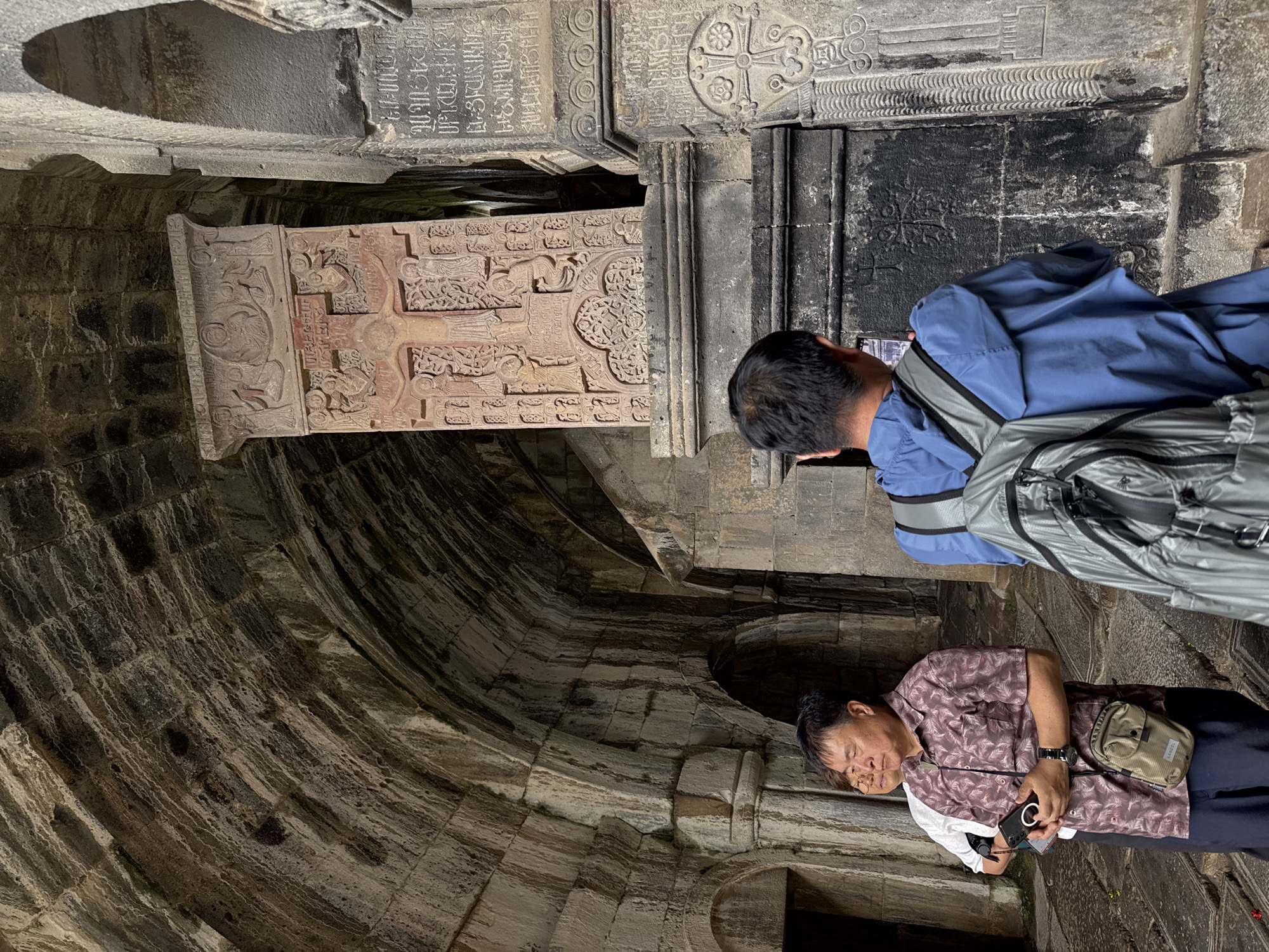 The image captures a scene inside the Haghpat Monastery complex in Haghpat, Armenia. In the foreground, two men are visible. One man, facing away from the camera and wearing a blue jacket and a gray backpack, is holding a phone, appearing to take a picture. To his left, another man in a patterned short-sleeved shirt and dark pants is holding a phone and looking to his right. Behind these two men, another individual, partially obscured, stands in the background.
The environment is characterized by ancient stone architecture. A prominent stone khachkar, intricately carved with religious imagery and Armenian inscriptions, stands in the center of the frame. To the right of the khachkar, another carved stone pillar with inscriptions and decorative motifs is visible. The ceiling of the structure is a ribbed stone vault, showing signs of age and wear. Arched passageways and stone walls surround the individuals, contributing to the historical and religious atmosphere of the location. The lighting suggests an overcast day or an indoor setting with ambient light.
