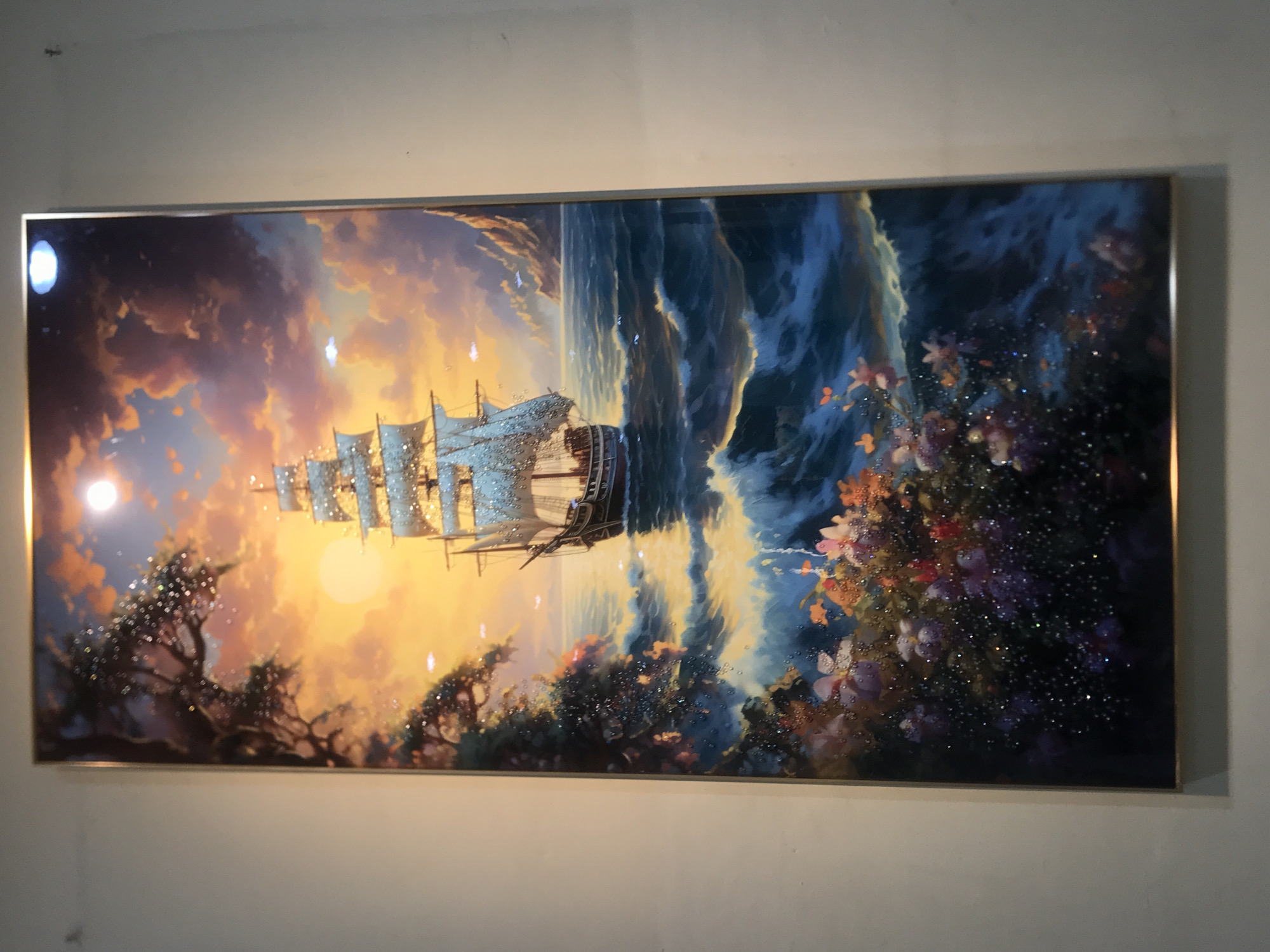 A framed rectangular painting hangs on a light grey wall. The painting depicts a sailing ship at sea during a sunset or sunrise. The left side of the painting shows the sky with shades of orange, yellow, and purple. Silhouetted trees and foliage are at the bottom left. The ship is near the center, with sails extended, appearing to sail towards a bright light. The right side of the painting shows a turbulent ocean with waves crashing and dark blue and white hues. Flowers are visible near the bottom right. The frame is thin and metallic. The location is City of Medan, Indonesia.