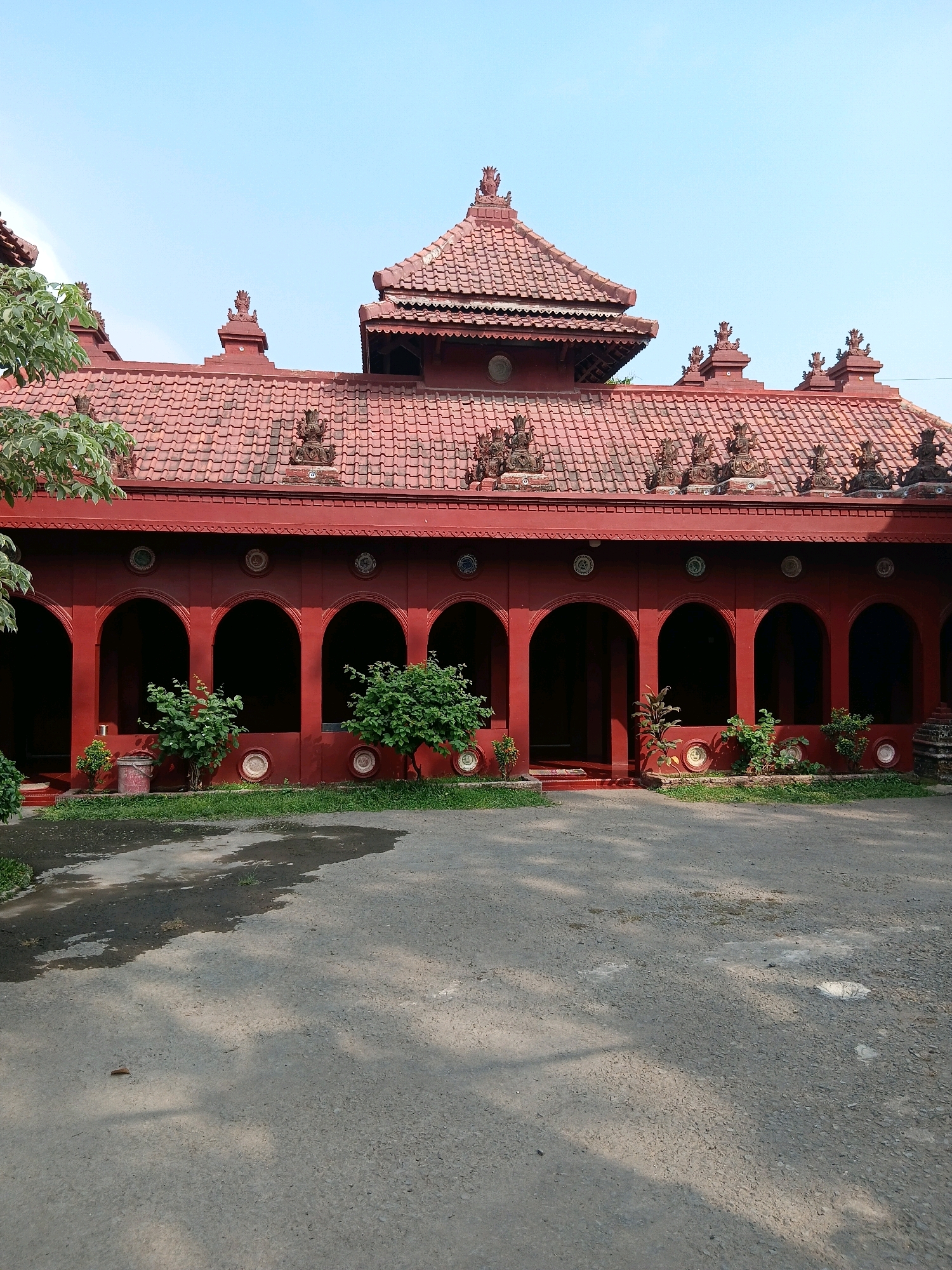 The image depicts the exterior of the Sang Cipta Rasa Grand Mosque, a significant historical building located in Cirebon, West Java, Indonesia. The mosque features traditional Javanese architectural elements, characterized by its red-tiled roof with ornate finials and intricately carved decorations along the eaves. A series of arched doorways or openings form a covered walkway along the front facade of the building, with several small trees and decorative elements placed along the walkway. The ground in front of the mosque is paved with asphalt, showing some patches of moisture and shadow from the surrounding trees. The sky above is clear and blue. There are no people visible in the frame.