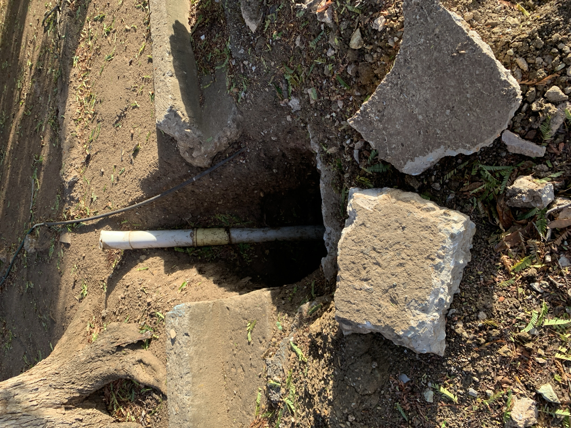 In the city of Santa Ana, Venezuela, a shallow excavation reveals a section of white PVC pipe running vertically into the ground. The pipe has a yellow band around its middle and appears to be connected to a black cable or hose extending upwards and to the right. The area around the excavation is covered in dirt and scattered with dry leaves and small green plants. Two large, broken pieces of concrete lie on the ground near the pit, one in the foreground to the left and another to the right. A large tree trunk with exposed roots is visible on the left side of the image. The scene appears to be outdoors, with sunlight casting shadows on the ground.