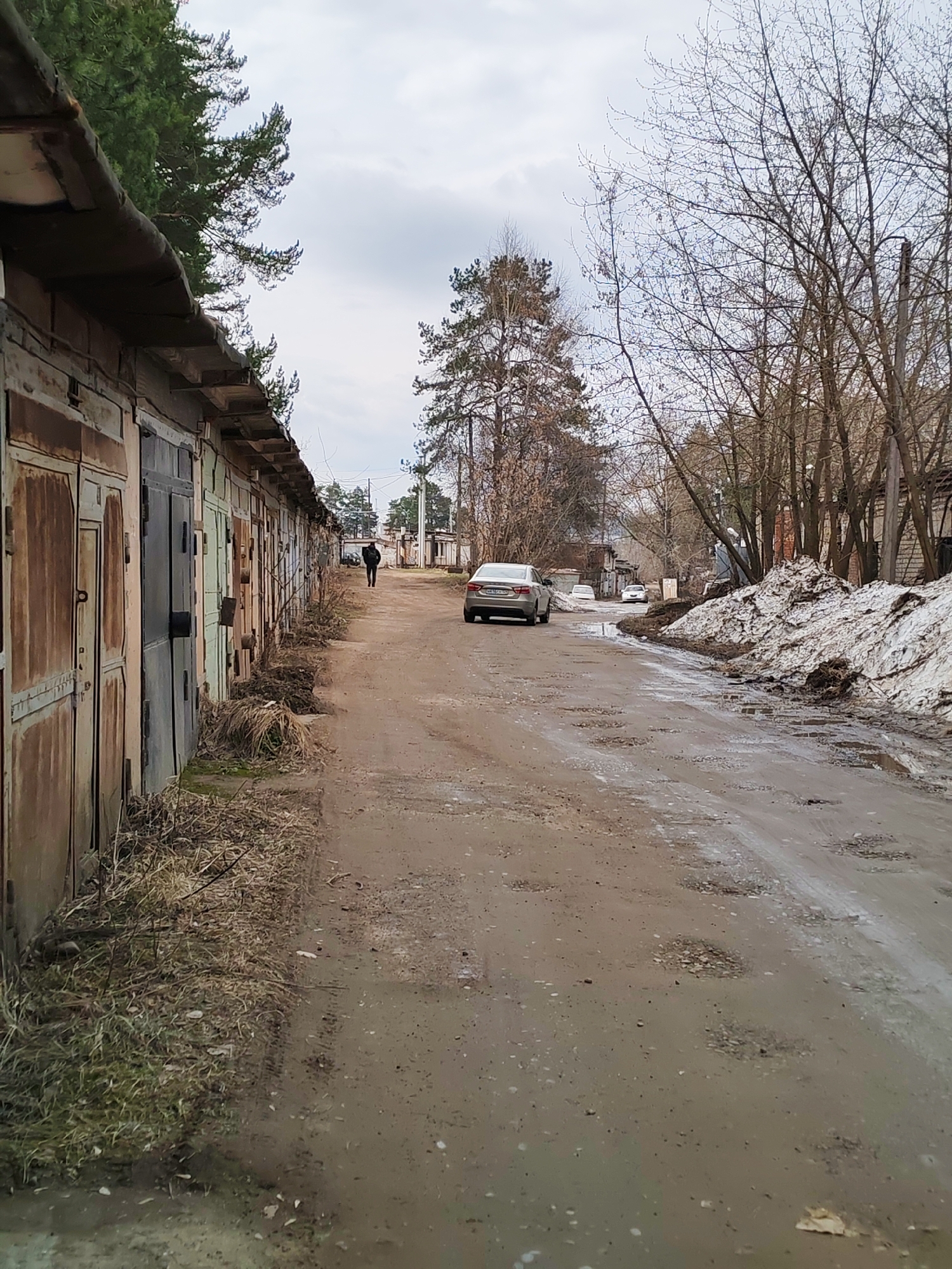 The image depicts a street in what appears to be an industrial or semi-urban area of Chaikovsky, Russia. On the left side of the frame is a long row of weathered, adjacent garage doors. The doors exhibit varying degrees of rust and discoloration, suggesting age and lack of maintenance. Sparse, dead grass and debris accumulate along the base of the garages.
A man in dark clothing is walking down the street away from the viewer. A silver sedan is parked on the right side of the road facing towards the viewer. The road is unpaved and muddy, with numerous potholes and puddles. A large pile of melting snow sits to the right of the car. Bare trees line the right side of the road, their branches devoid of leaves, indicating it is likely early spring or late autumn. The sky is overcast, lending a somber tone to the scene. In the distance, other cars are parked along the street. The scene suggests a transition period between seasons, with remnants of winter still present in a landscape showing signs of thawing.