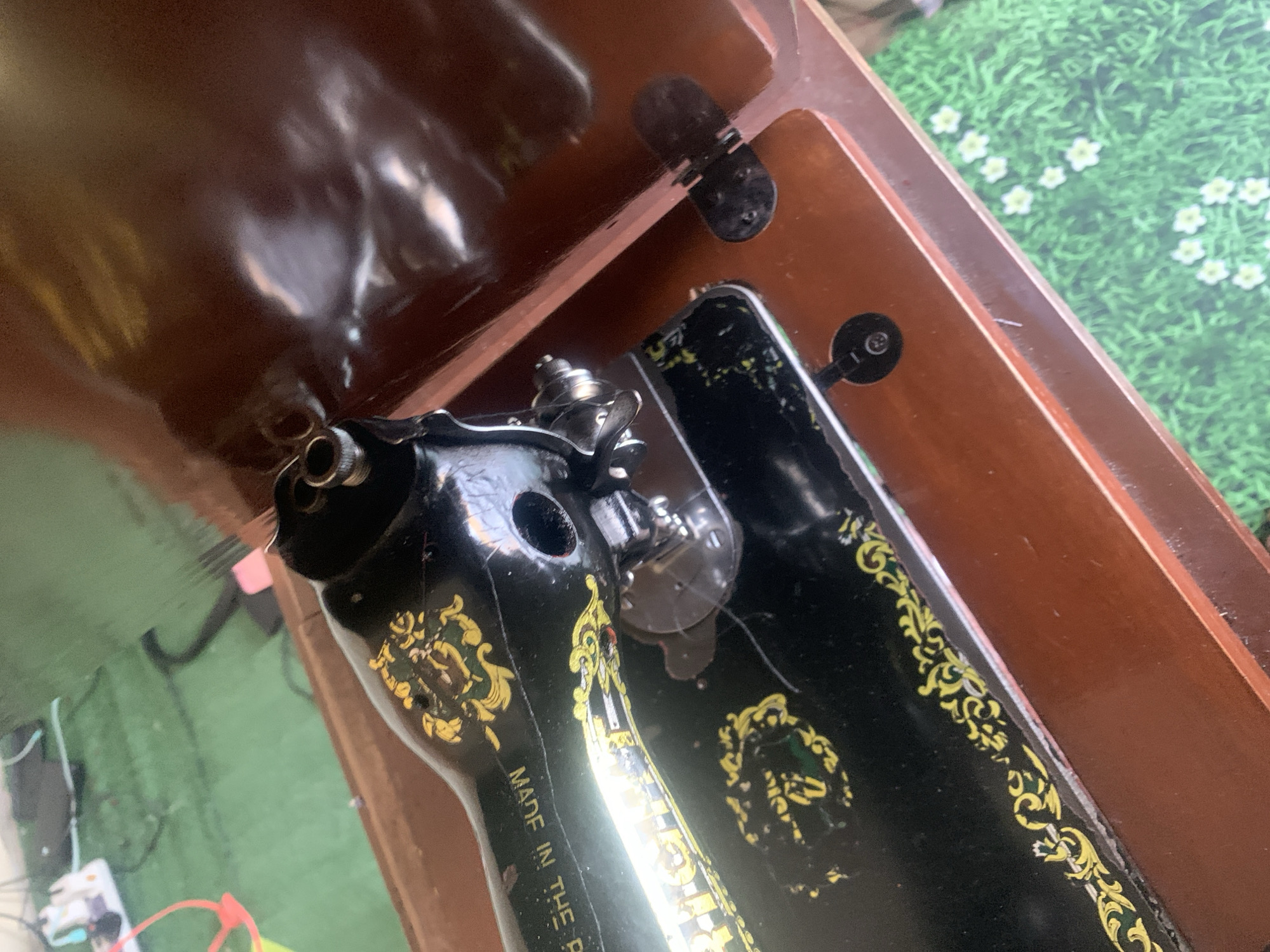 A vintage black sewing machine with gold ornamentation is shown in the image. The sewing machine is housed within a wooden case, the lid of which is open and angled to the right. The sewing machine's deck is adorned with intricate floral patterns. The image is captured from a close-up perspective, focusing on the sewing machine and its immediate surroundings. The background is a green surface with a pattern of small white flowers. The scene does not provide any specific information about the city of Jauro Yinu, Nigeria, or any people present.