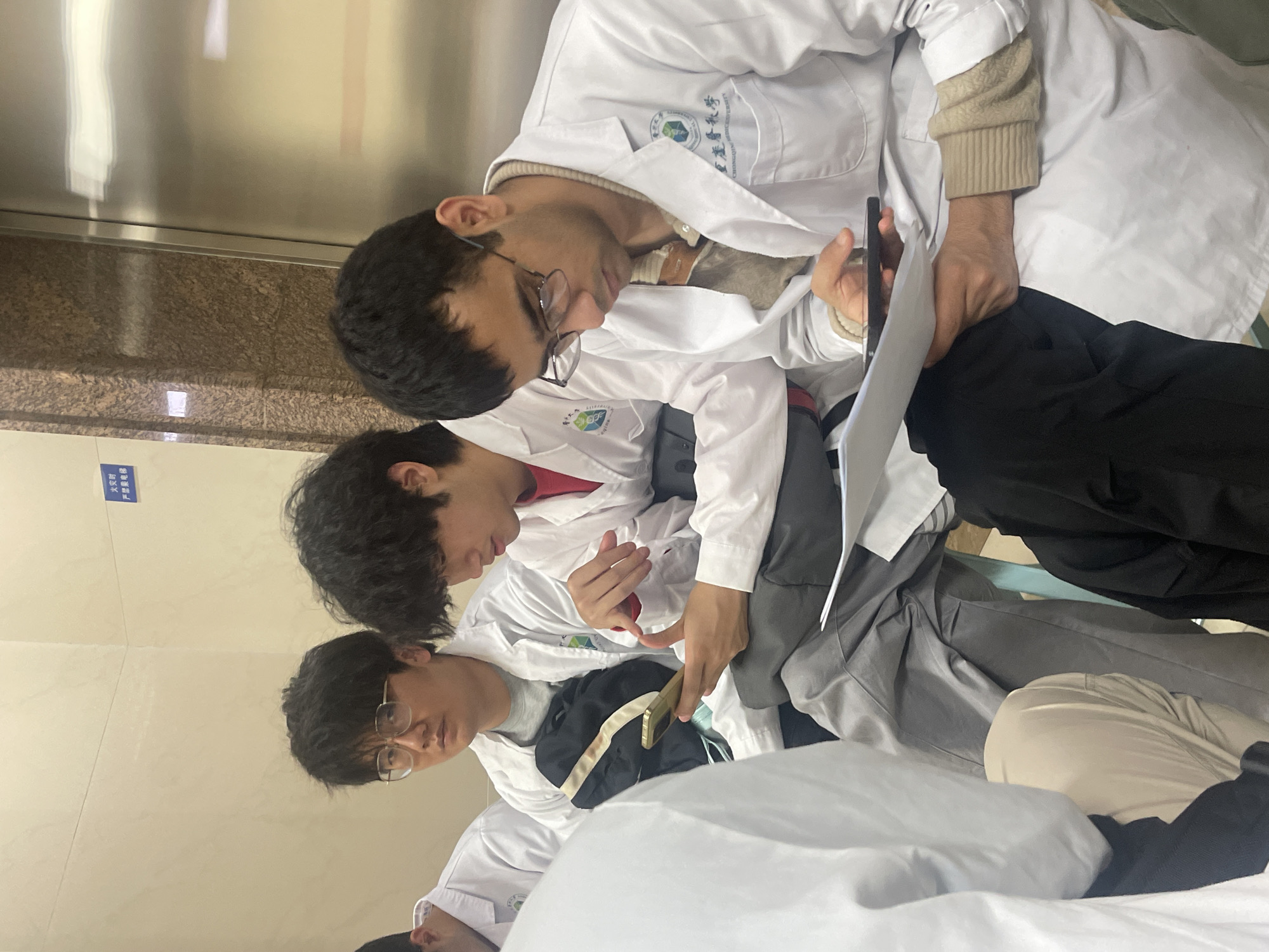 The scene depicts four males seated in an indoor hallway or waiting area within a facility associated with Chongqing Medical University, located in Yuzhong District, Chongqing, China. All visible individuals are wearing white lab coats over casual clothing.
From left to right:
1. A male, partially visible, wearing a white lab coat over a red undershirt.
2. An East Asian male with dark hair and spectacles, looking forward with a neutral expression. He wears a grey undershirt under his lab coat, and a black bag rests on his lap.
3. An East Asian male with dark, wavy hair, intently viewing a gold-colored smartphone held horizontally in both hands. He wears a red undershirt under his lab coat and grey trousers. His lab coat displays a partially obscured logo.
4. A South Asian male with dark hair and spectacles, looking down at a black smartphone or tablet held in his left hand, with white documents resting on his lap and held by his right hand. He wears a beige patterned sweater under his lab coat and black trousers. His lab coat clearly displays the "CHONGQING MEDICAL UNIVERSITY" logo in English and Chinese characters.
The background features a plain light-colored wall with a blue sign mounted on it, displaying Chinese characters "火灾时 严禁乘电梯" (meaning "In case of fire, elevators are strictly prohibited"). To the right, a reflective metallic surface, consistent with an elevator door, is visible, flanked by a brown speckled marble-like column. The individuals are engaged with personal devices or documents; no direct inter-personal interaction is observed.