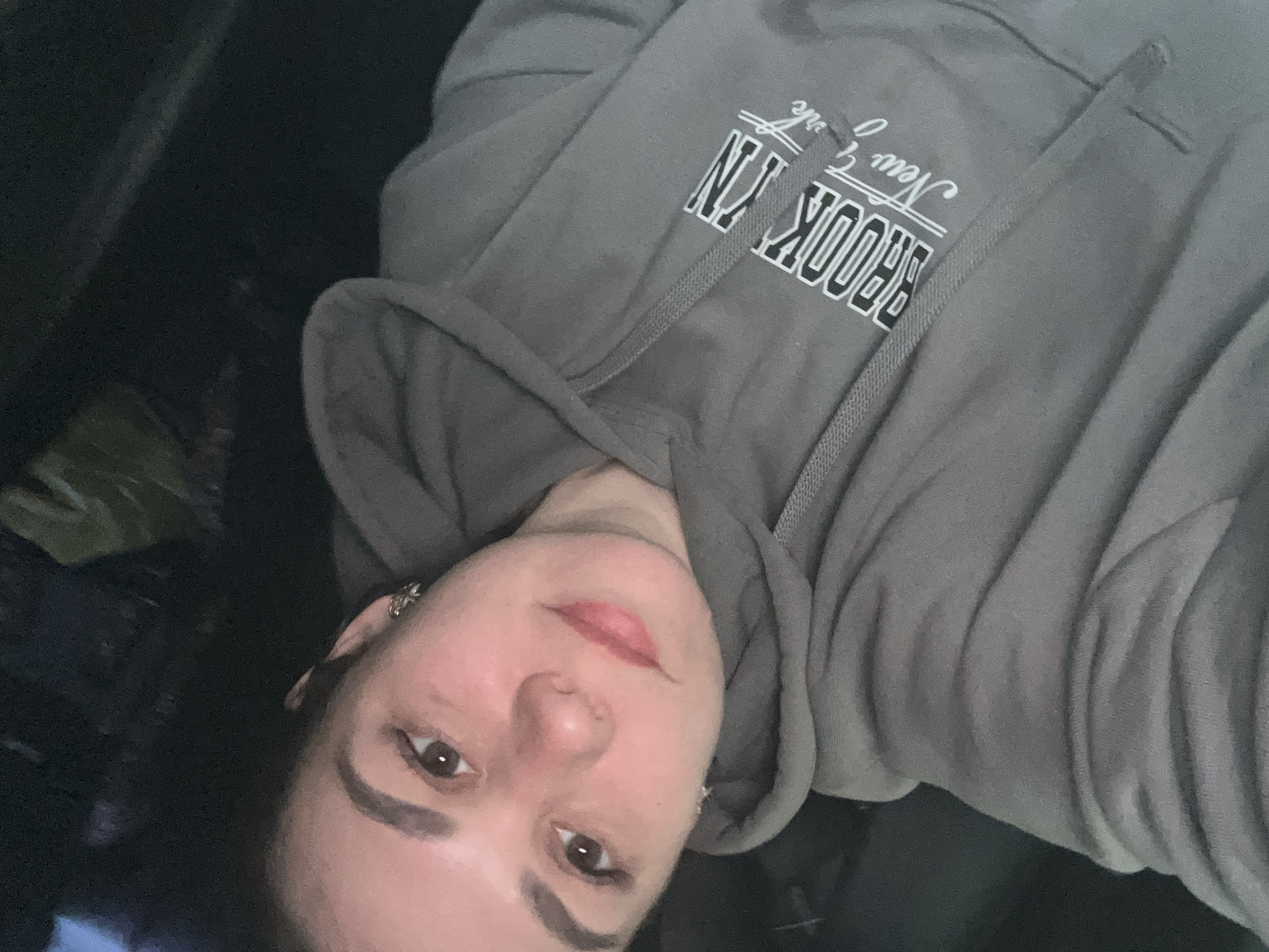 A person wearing a gray hooded sweatshirt with "BROOKLYN New York" printed on the chest is lying down. The person is facing the camera with a slight upward gaze. The background is dark and indistinct. There are no discernible features indicating the location is in the city of Chaikovsky, Russia.