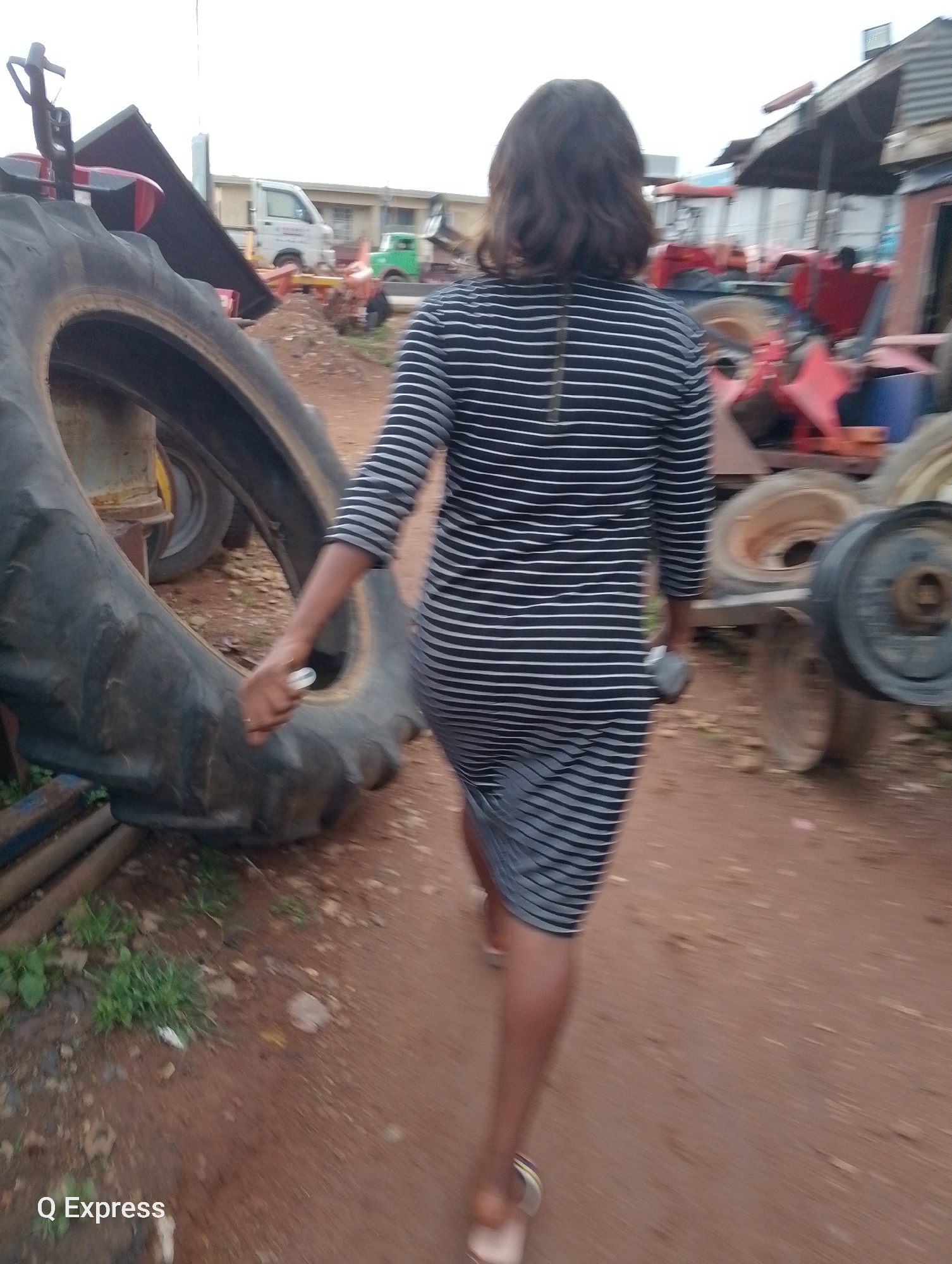 The image shows a woman from behind, wearing a black and white striped dress, walking on a dirt path in what appears to be a scrapyard or mechanic's workshop. In the foreground, a large, worn tractor tire is visible. Several other tractor tires and parts are scattered around the scene, along with what looks like a red tractor in the background and some buildings. The environment suggests an outdoor industrial or salvage area in Jos, Nigeria, indicated by the watermark "Q Express" in the lower left corner. The lighting suggests it is daytime.