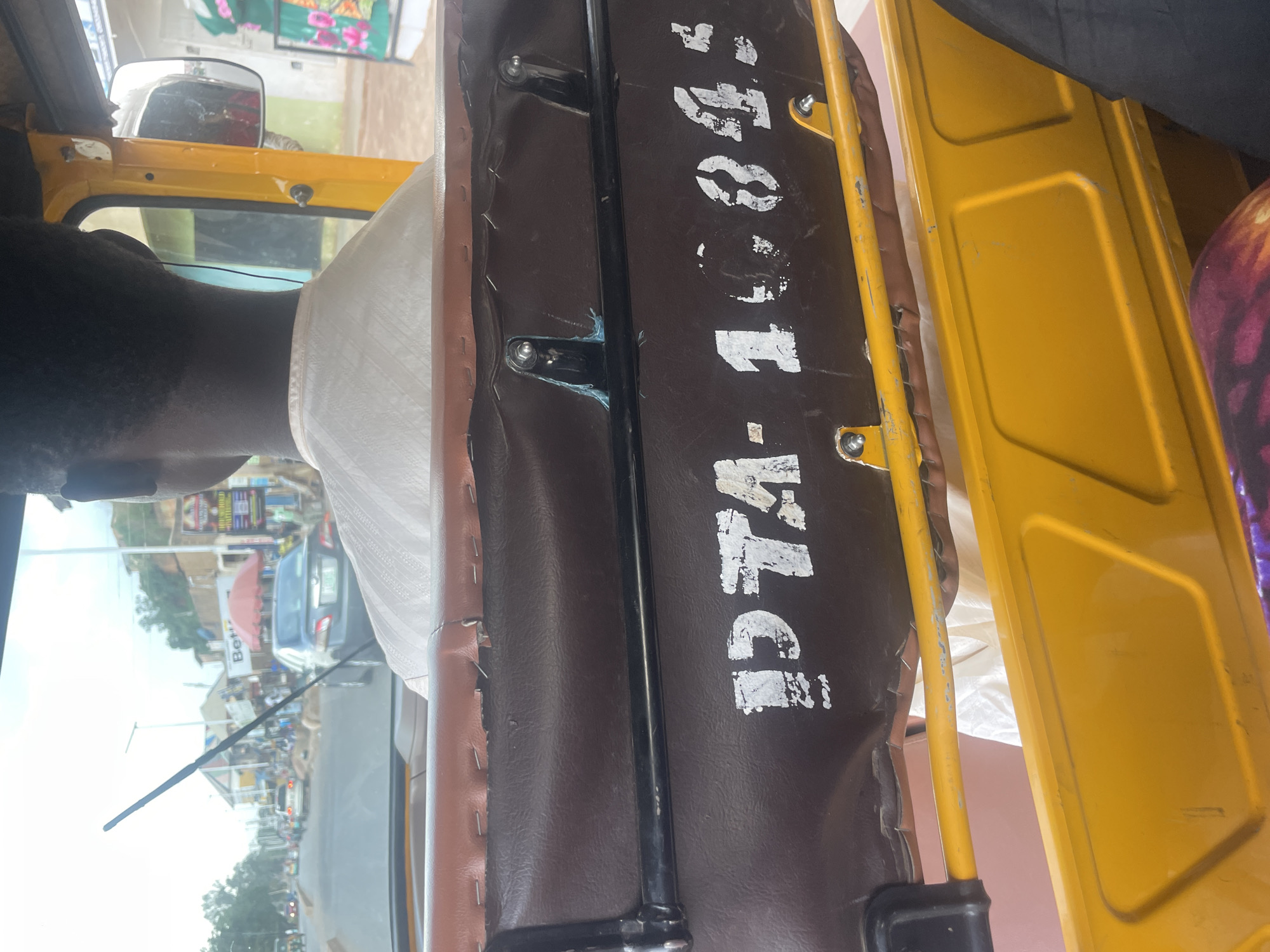 The image captures a view from the interior of a yellow auto-rickshaw, possibly in Barakin Lerawi, Nigeria. The focus is on the back of a person wearing a white garment, whose head and shoulders are visible from behind. The auto-rickshaw has a brown seat in the foreground with white lettering that reads "IDTA-10945". The outdoor environment outside the vehicle appears to be a street scene with buildings, other vehicles, and people. The sky is overcast, suggesting a cloudy day. The overall impression is of a candid shot taken while traveling in the auto-rickshaw.